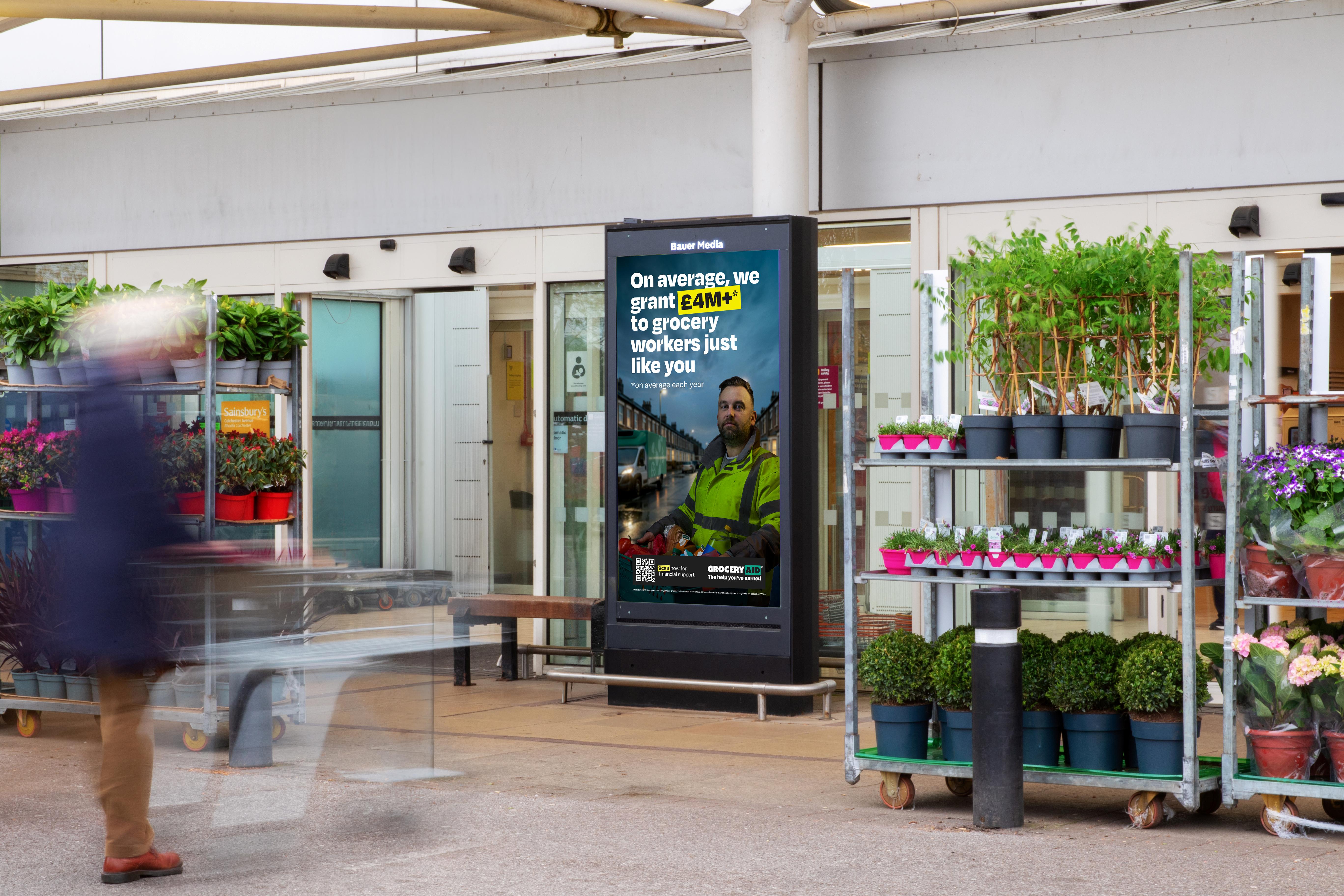 A blurred person walks past the entrance of a small shop, with colorful displays and potted plants arranged outside on the pavement.