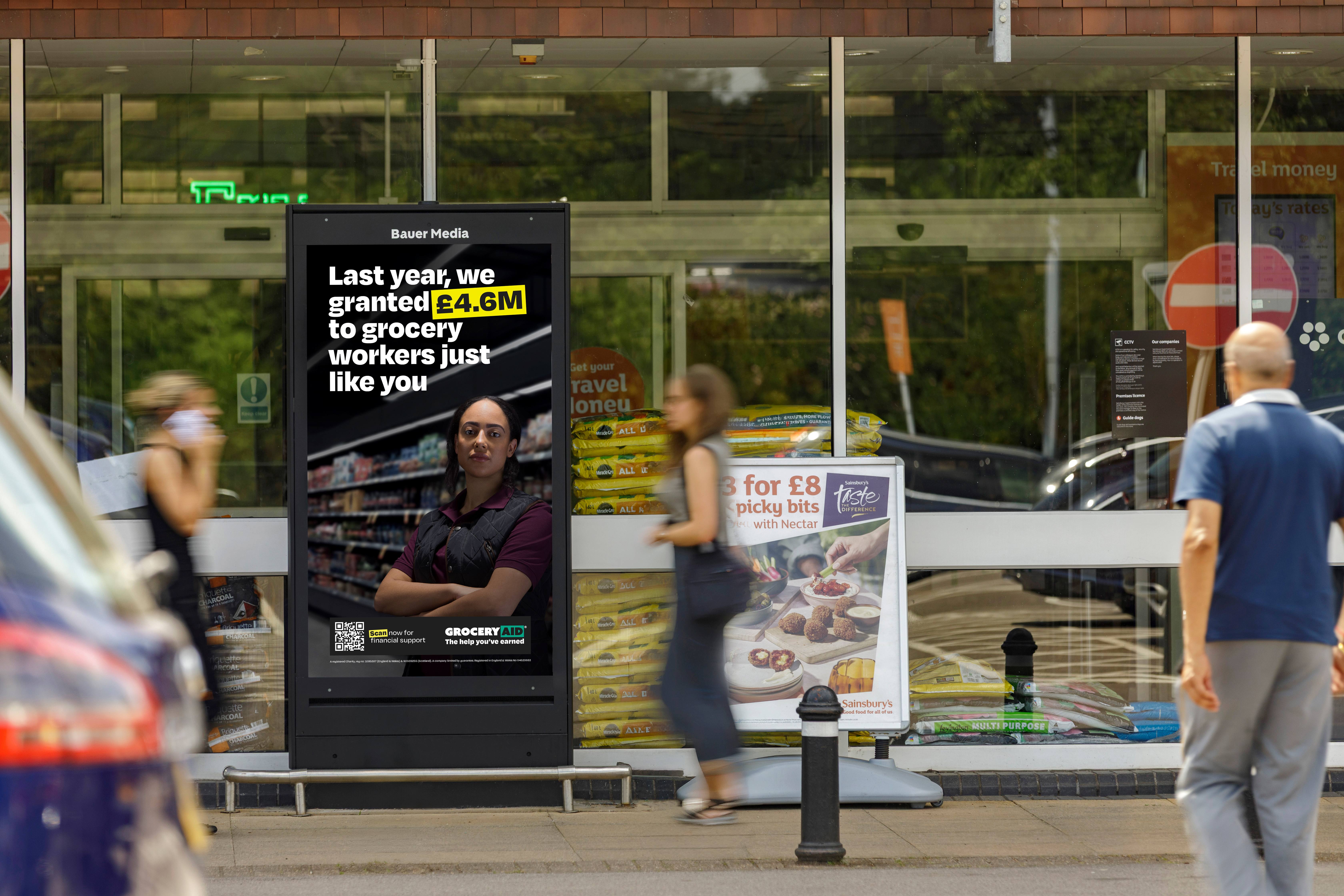 A supermarket entrance hosts a digital billboard highlighting financial support available to grocery workers as shoppers pass by on a busy day.
