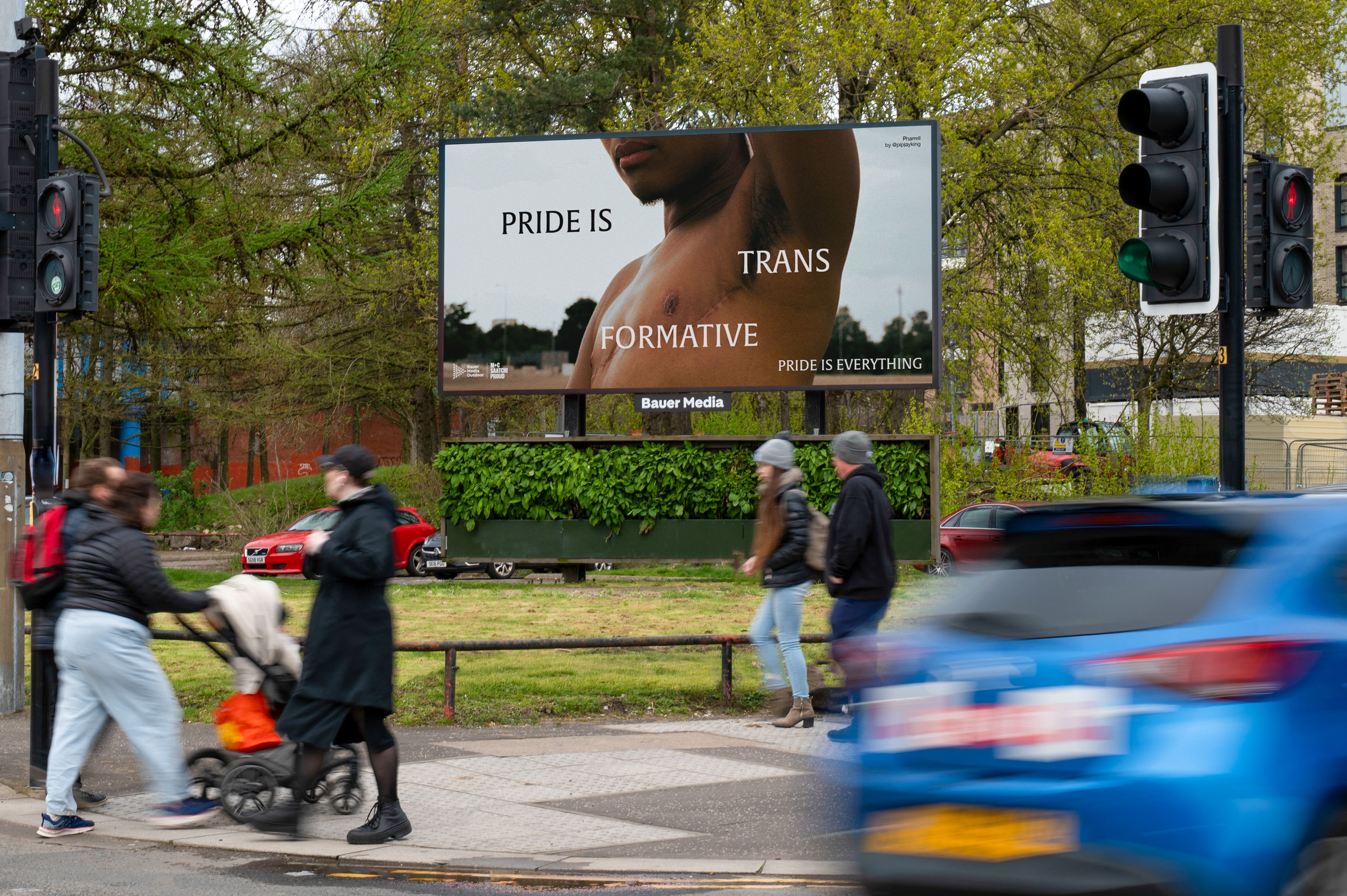 A city street scene with pedestrians and a blue car passing a large Bauer Media billboard. The billboard shows a close-up of a person’s top surgery scar with the text “Pride is transformative” and “Pride is everything,” with the word “trans” highlighted.