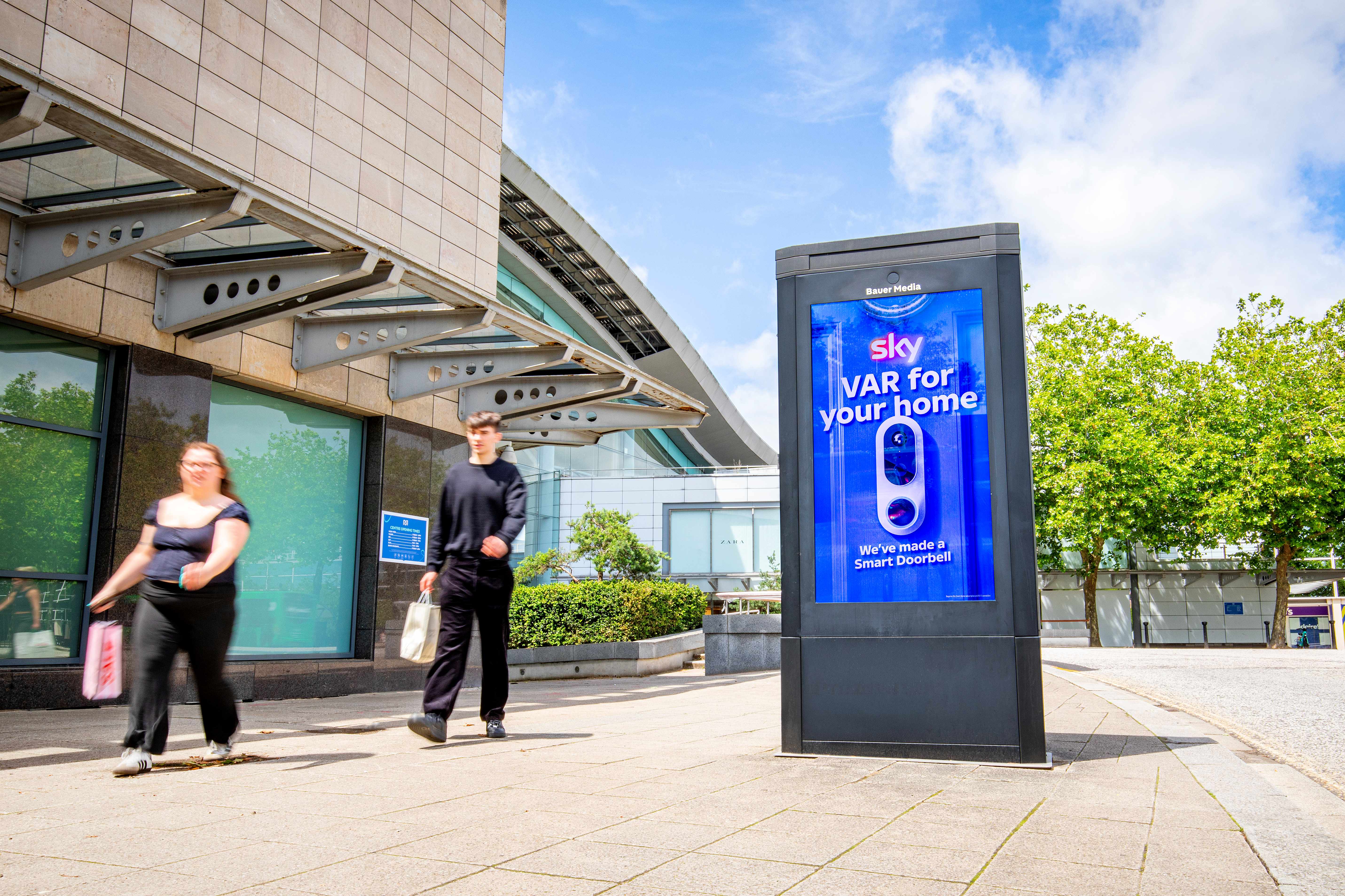 A digital advertising display outside a modern building shows a Sky Smart Doorbell ad reading “VAR for your home,” while two pedestrians walk past on a sunny pavement in Milton Keynes