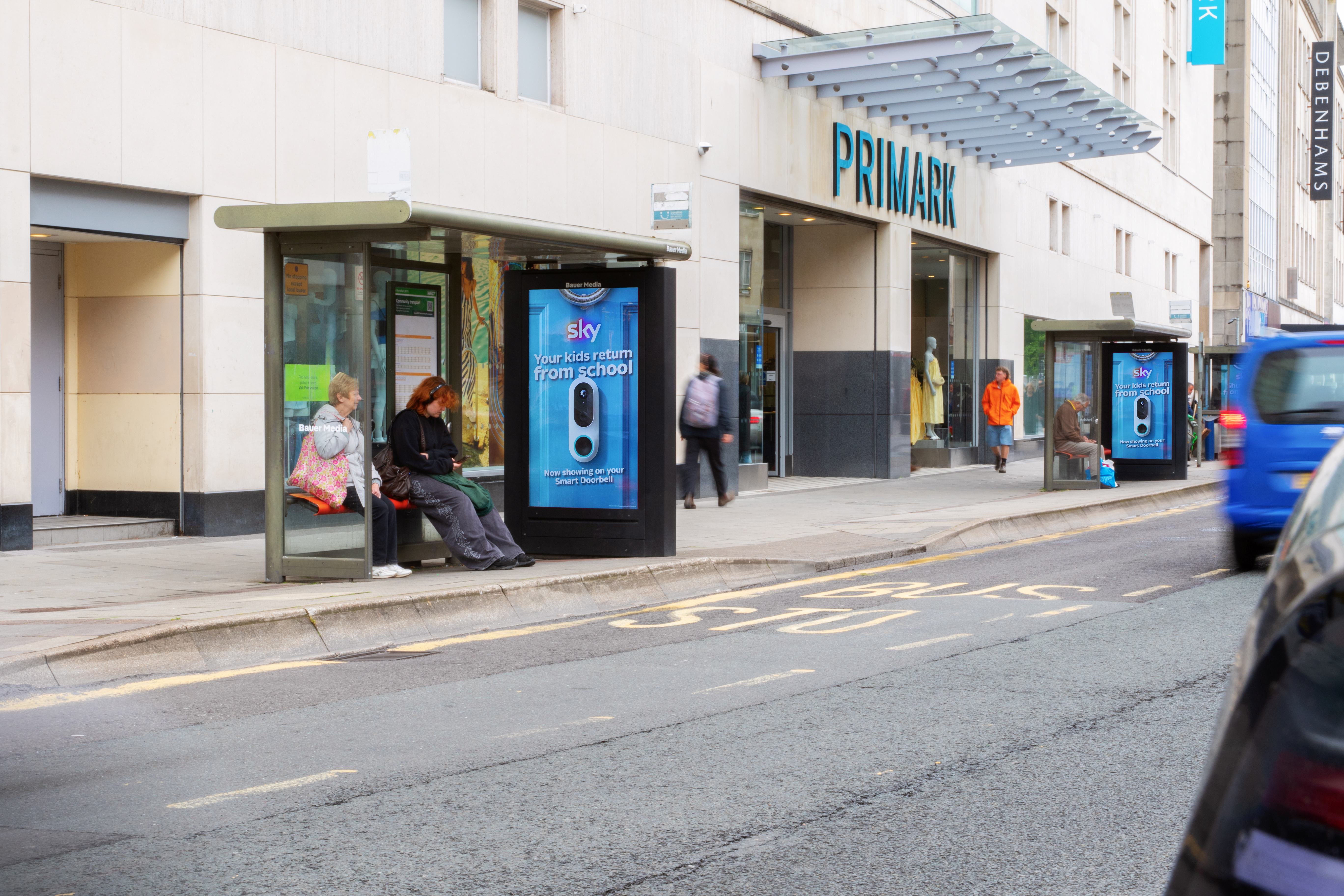 A Bristol street scene outside a Primark store, with people waiting at bus stops displaying digital Sky adverts. A blue van passes on the right-hand side of the road.