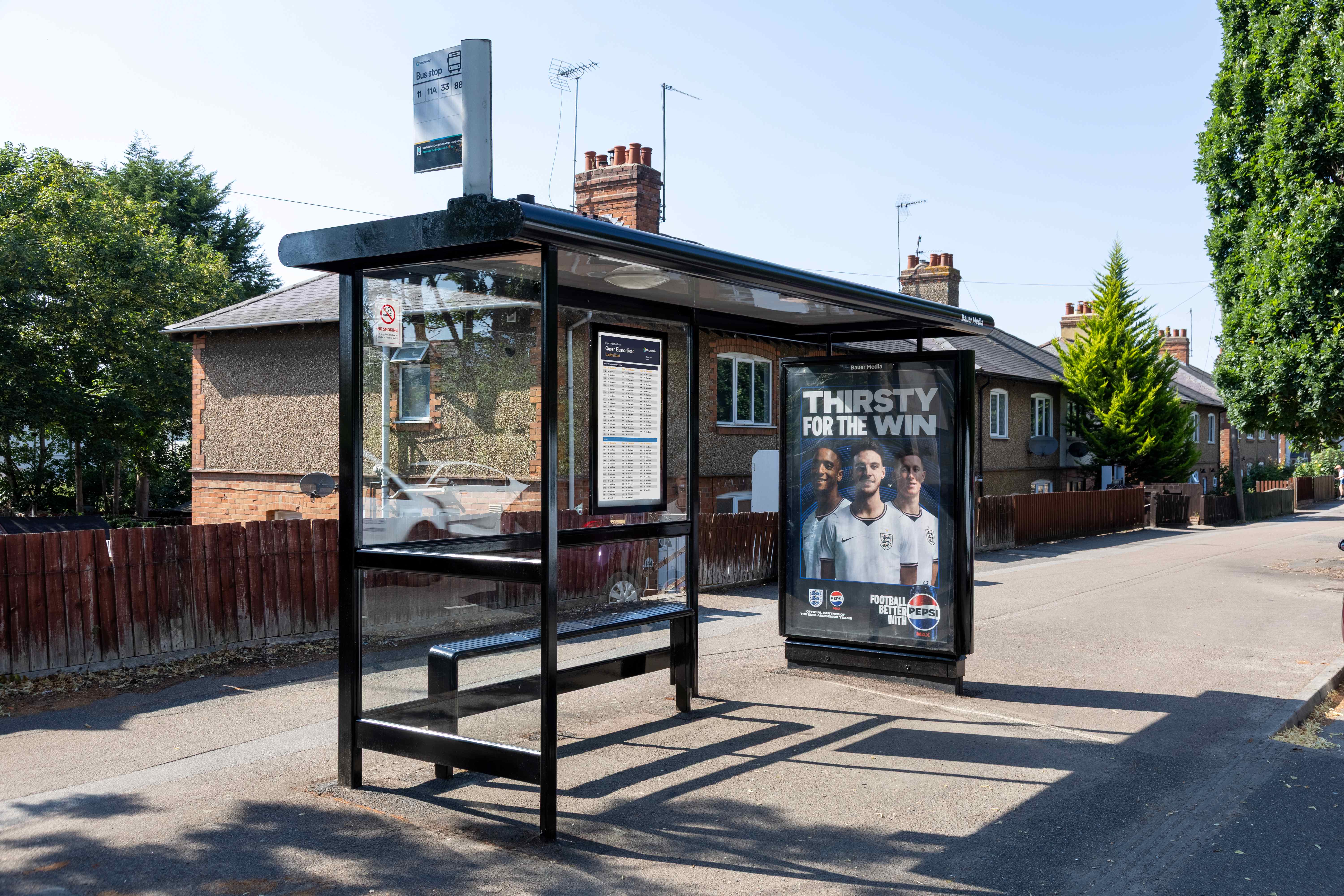A glass bus stop shelter on a sunny residential street, with a large advertising poster reading “Thirsty for the Win” displayed on a panel beside it.
