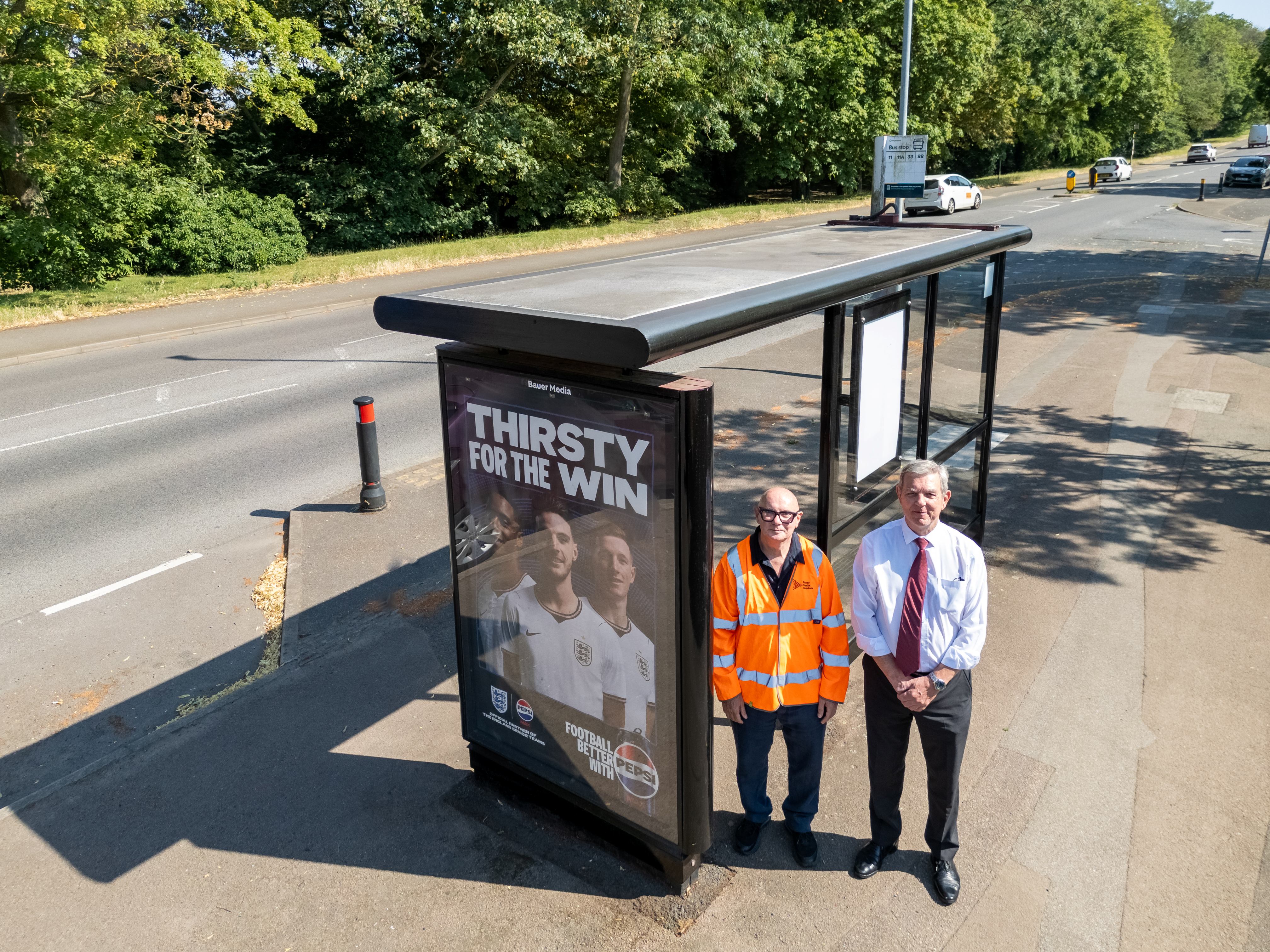 Two men stand beside a bus stop advertising panel displaying a “Thirsty for the Win” football-themed poster, next to a road lined with trees.#