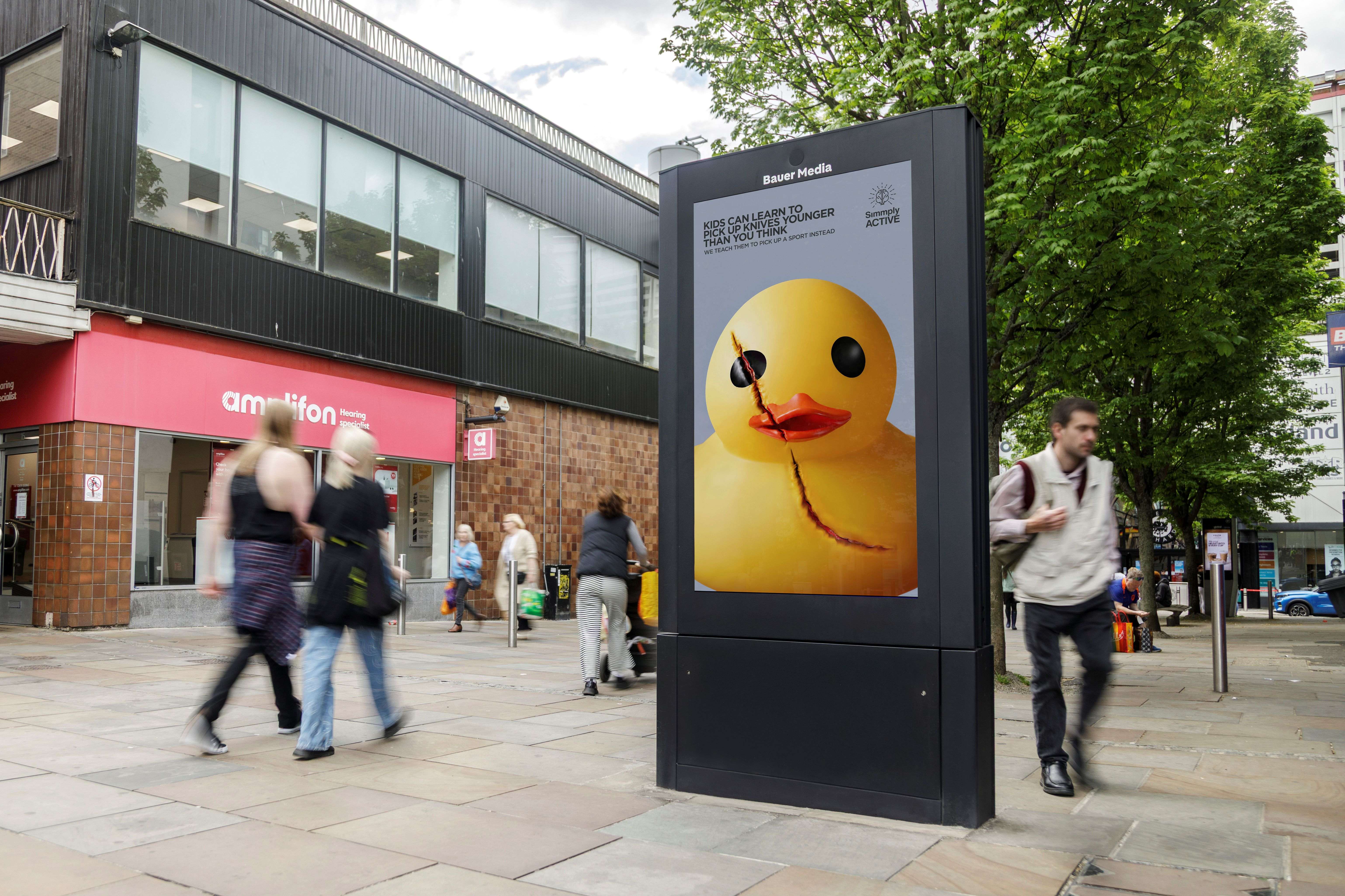 Bauer Media Outdoor adshel on a busy high street displays a campaign poster featuring a damaged yellow rubber duck and anti-knife crime messaging encouraging children to take up sport instead.