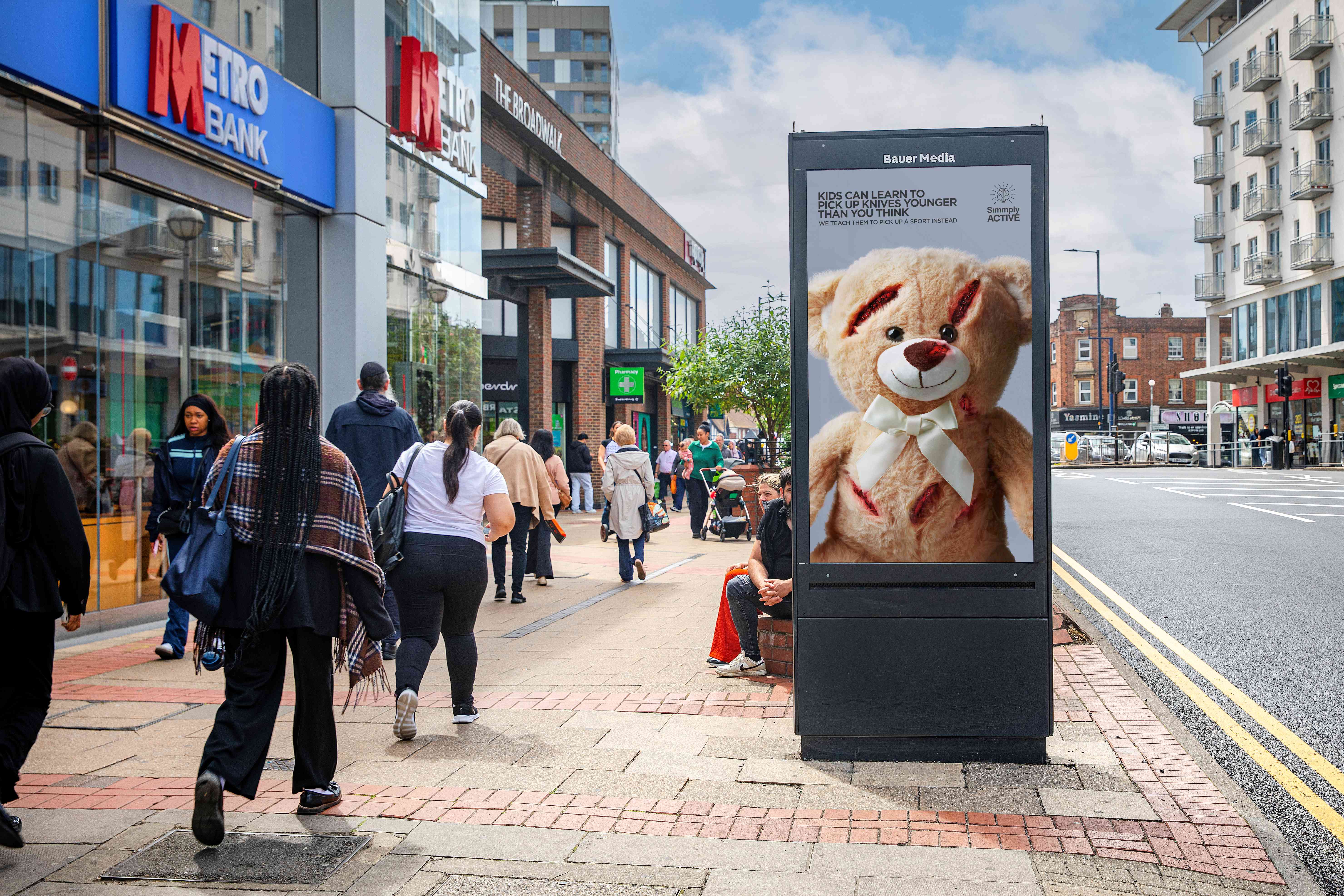  Bauer Media Outdoor Adshel on a city pavement showing a campaign poster of a teddy bear with cuts and the message: “Kids can learn to pick up knives younger than you think. We teach them to pick up a sport instead,” as pedestrians walk past nearby shops.