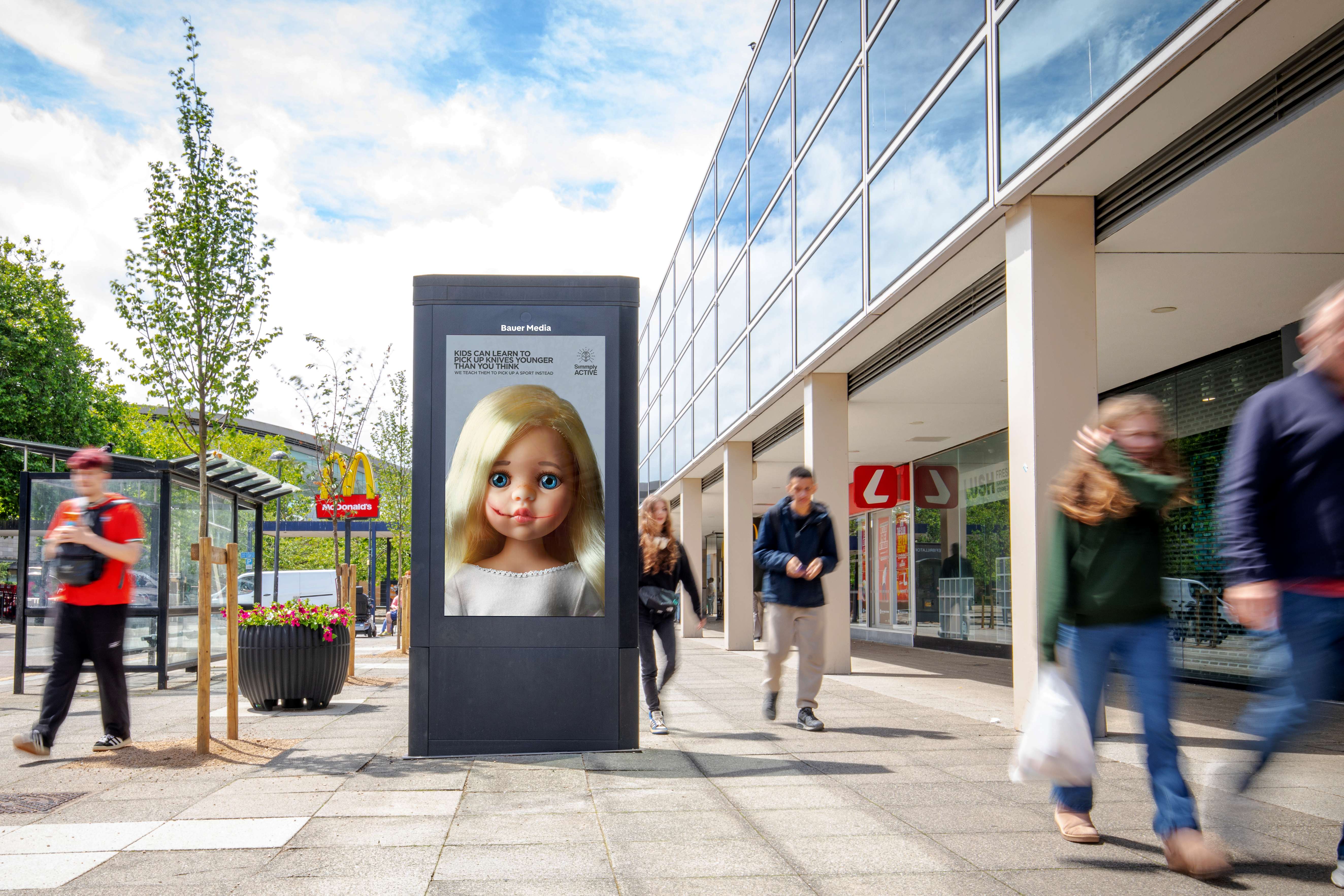 Street scene with a Bauer Media Outdoor adshel showing a campaign poster of a doll, alongside the message: “Kids can learn to pick up knives younger than you think. We teach them to pick up a sport instead.” as pedestrians walk past shops and a bus stop.