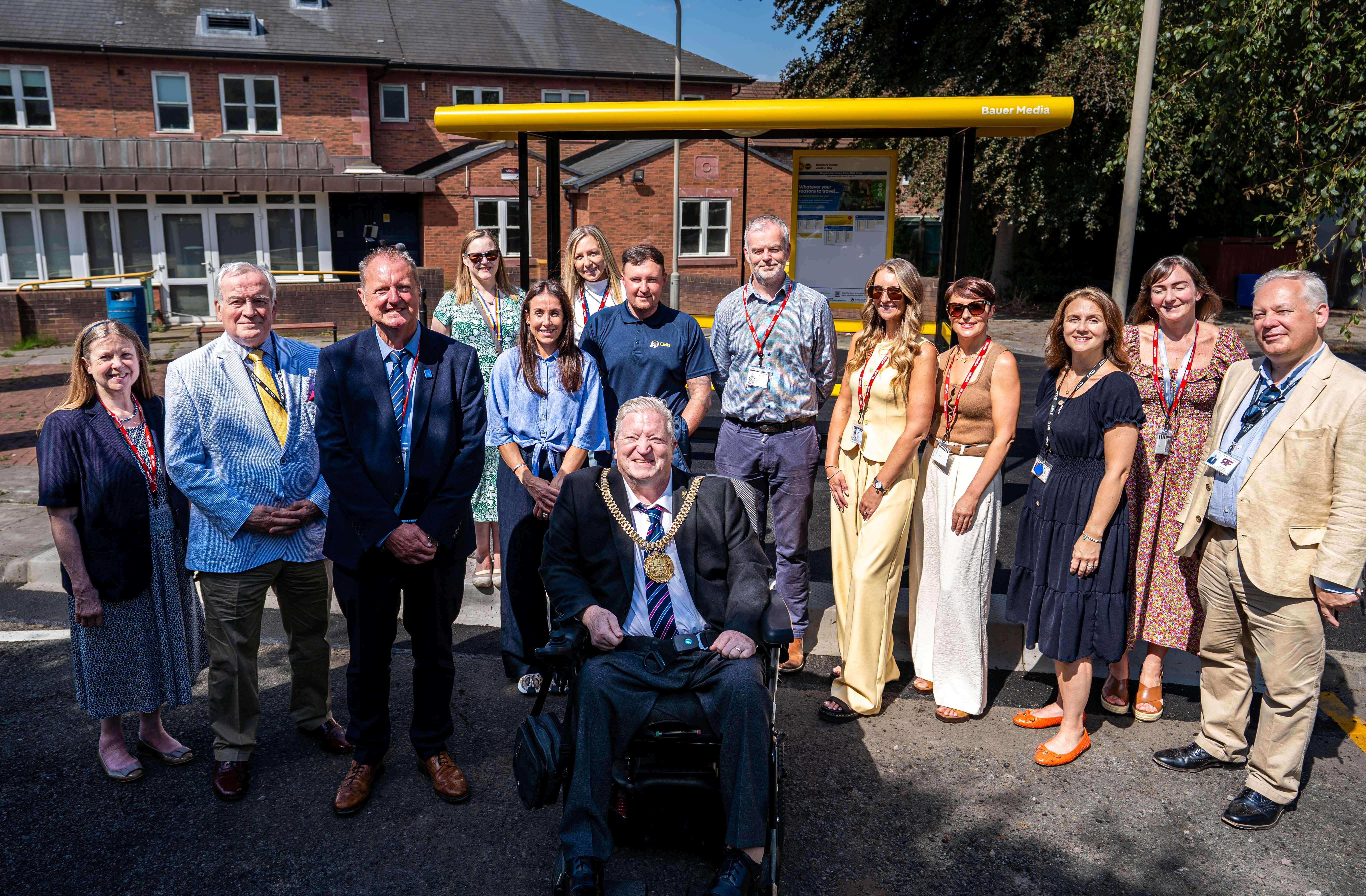 Bauer Media Outdoor staff with large group of staff and local representatives posing in front of a newly installed yellow Bauer Media-branded bus shelter outside a brick building