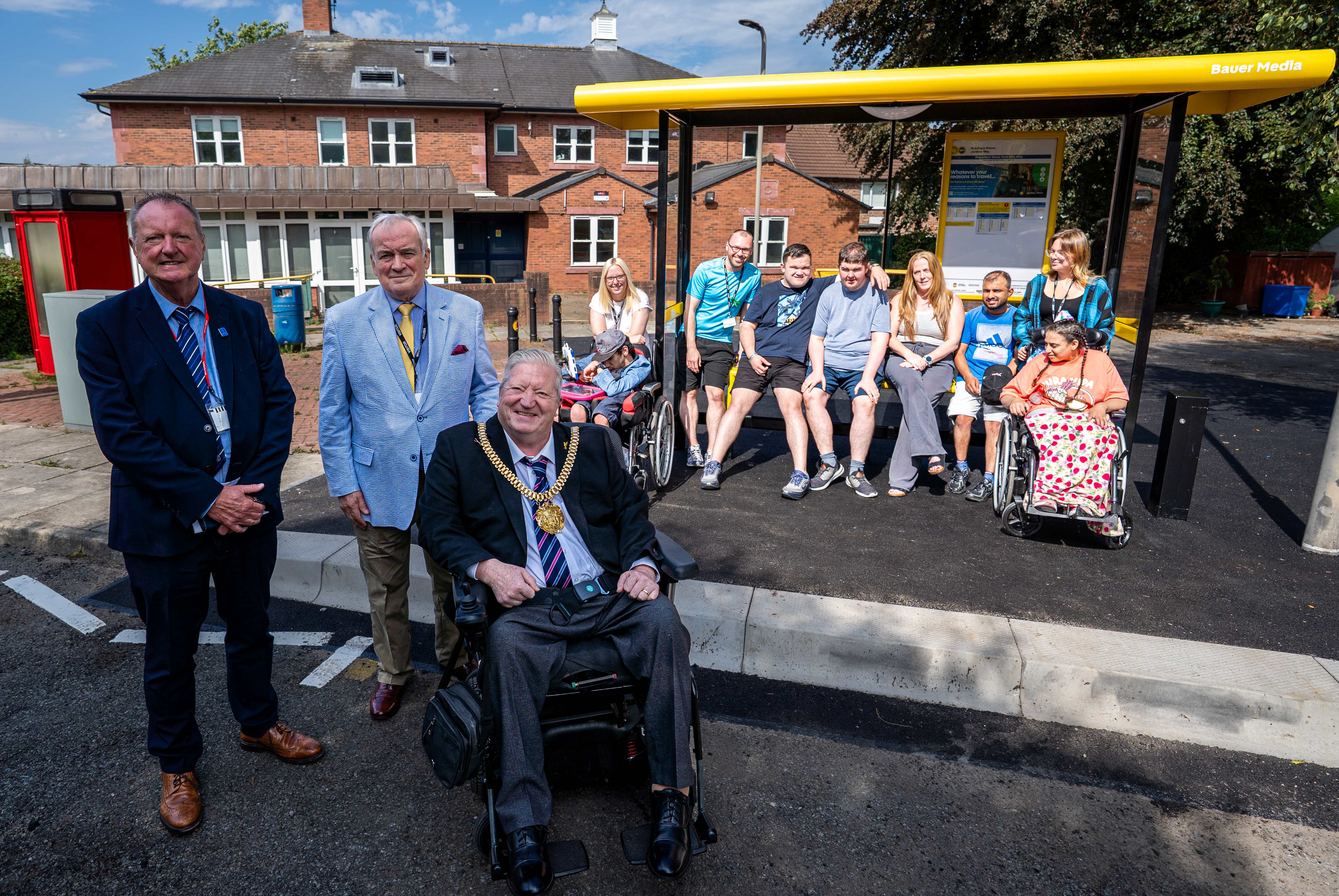 Group of people gathered beside a new yellow Bauer Media-branded bus shelter outside a brick building, with several people seated under the shelter and others standing nearby.