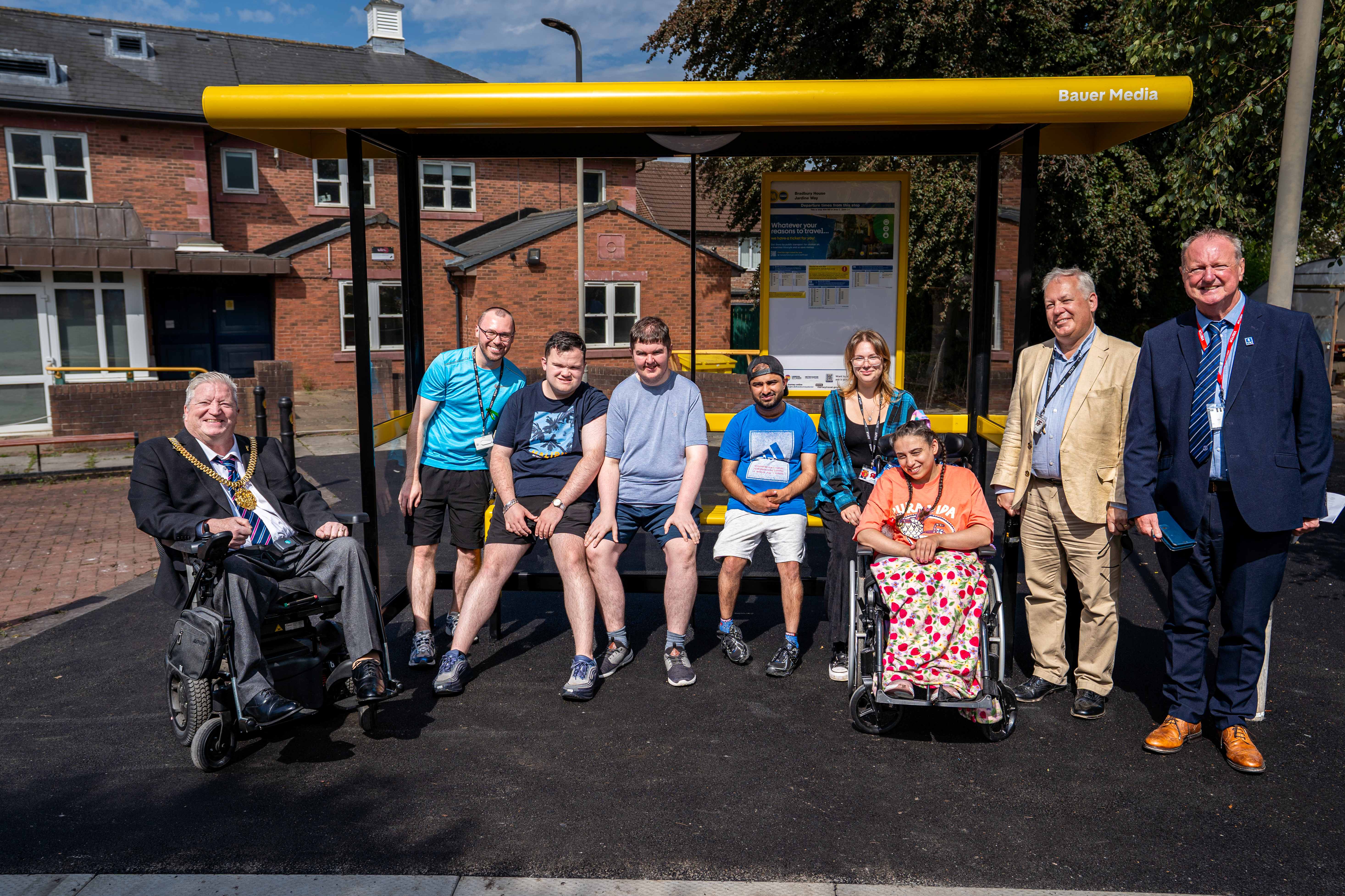Group photo of people standing and sitting in front of a yellow Bauer Media-branded bus shelter, including wheelchair users, staff and local representatives.