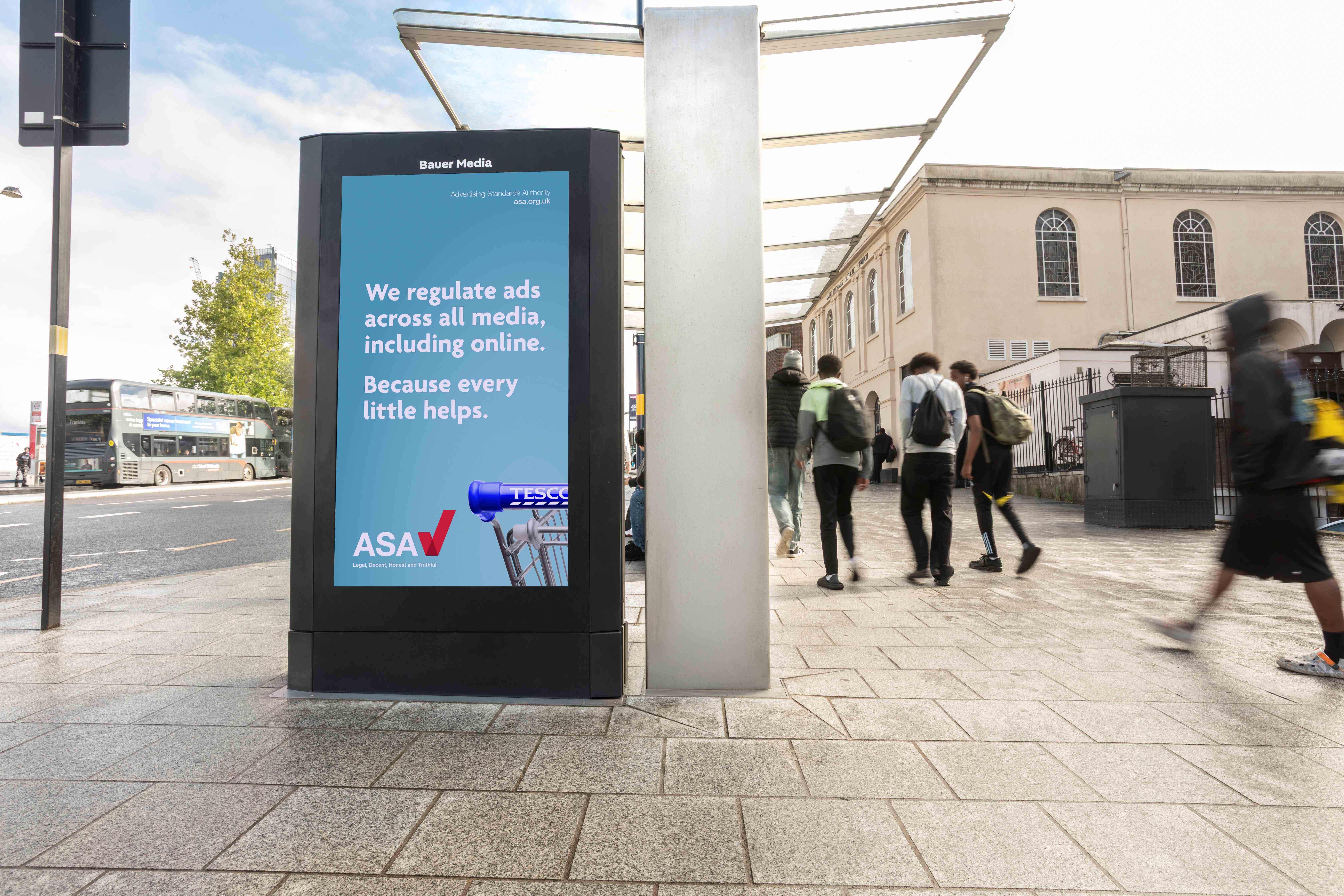 Bauer Media Outdoor Adshel Live advertising panel beside a busy pavement displays an ASA advert with the text: “We regulate ads across all media, including online. Because every little helps.” The ASA logo and a Tesco shopping trolley handle appear on the turquoise creative.