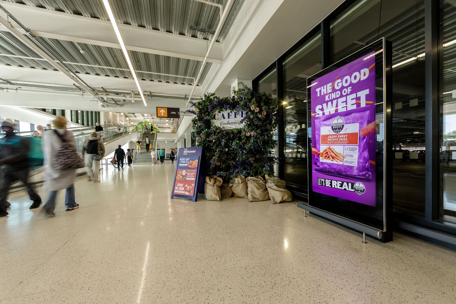 Digital screen inside a shopping centre displaying Strong Roots' "The Good Kind of Sweet" campaign for Crispy Sweet Potato Fries, positioned near a café and walkway