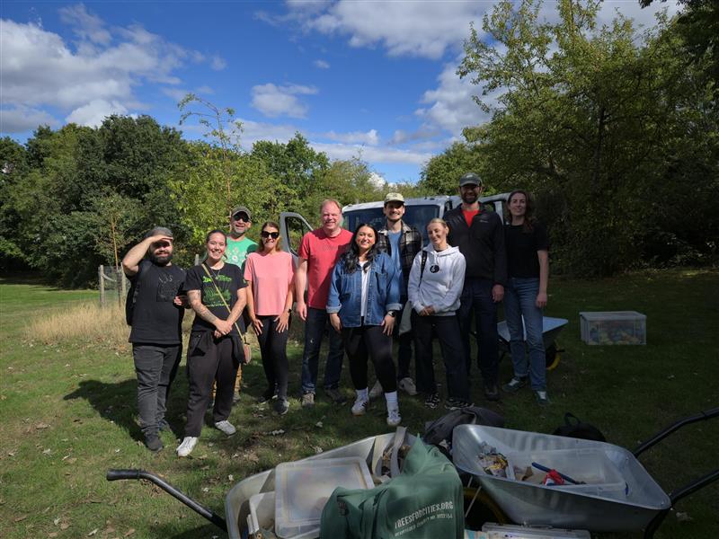 A group of Bauer Media Outdoor employees pose for a photo outdoors at Ravenor Park in Greenford after a volunteering session, with wheelbarrows of gardening tools and equipment in the foreground and trees under a blue sky in the background.