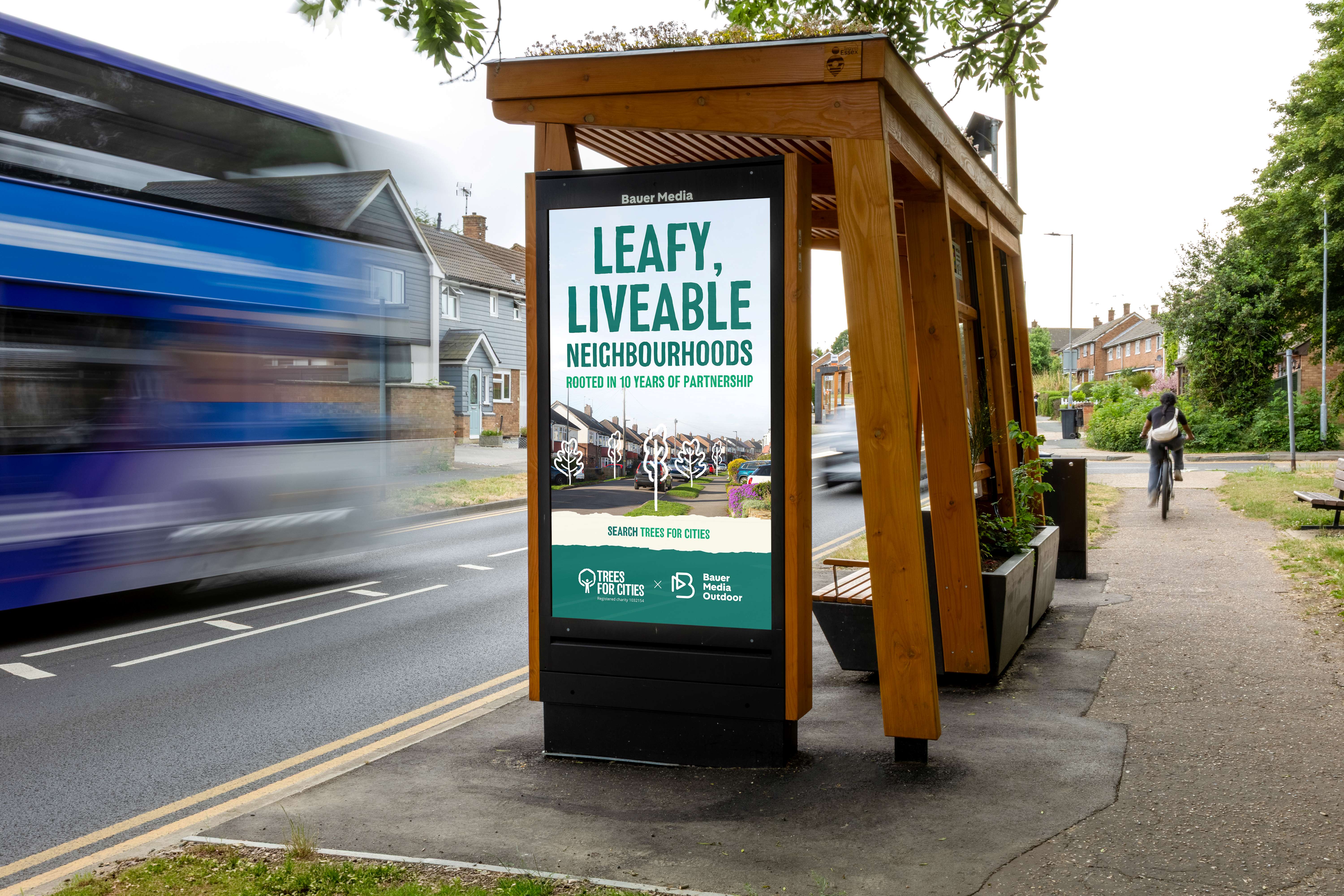 A wooden bus shelter with a green living roof on a residential street, featuring a digital OOH advertising screen that reads 'Leafy, Liveable Neighbourhoods – Rooted in 10 Years of Partnership' with the Trees for Cities and Bauer Media Outdoor logos. A bus passes by and a cyclist rides along the tree-lined pavement.