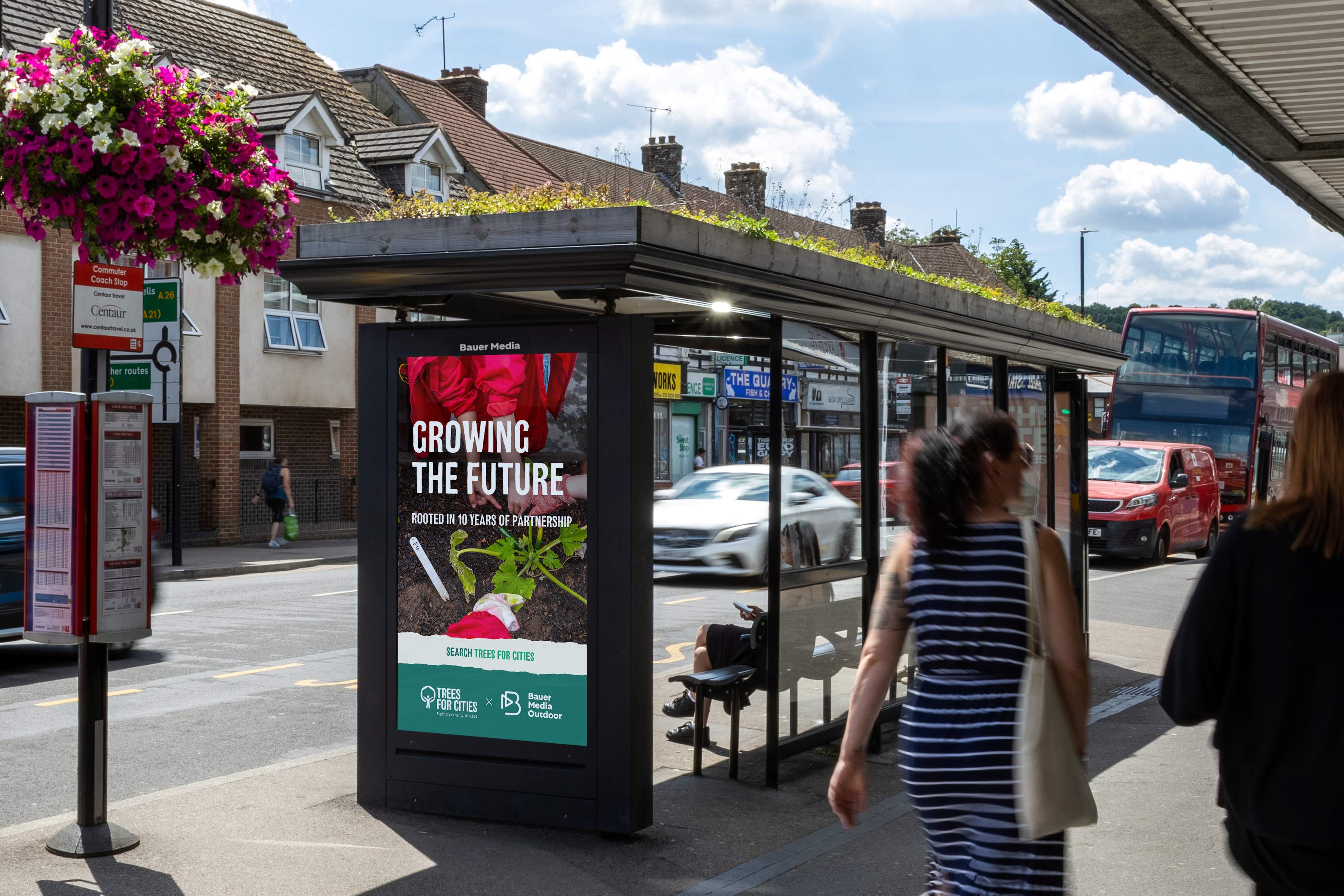 A bus shelter with a green living roof on a busy high street, displaying a digital advertising screen that reads 'Growing the Future – Rooted in 10 Years of Partnership' with the Trees for Cities and Bauer Media Outdoor logos. Pedestrians walk past as buses and cars travel along the road, with a hanging flower basket in the foreground.