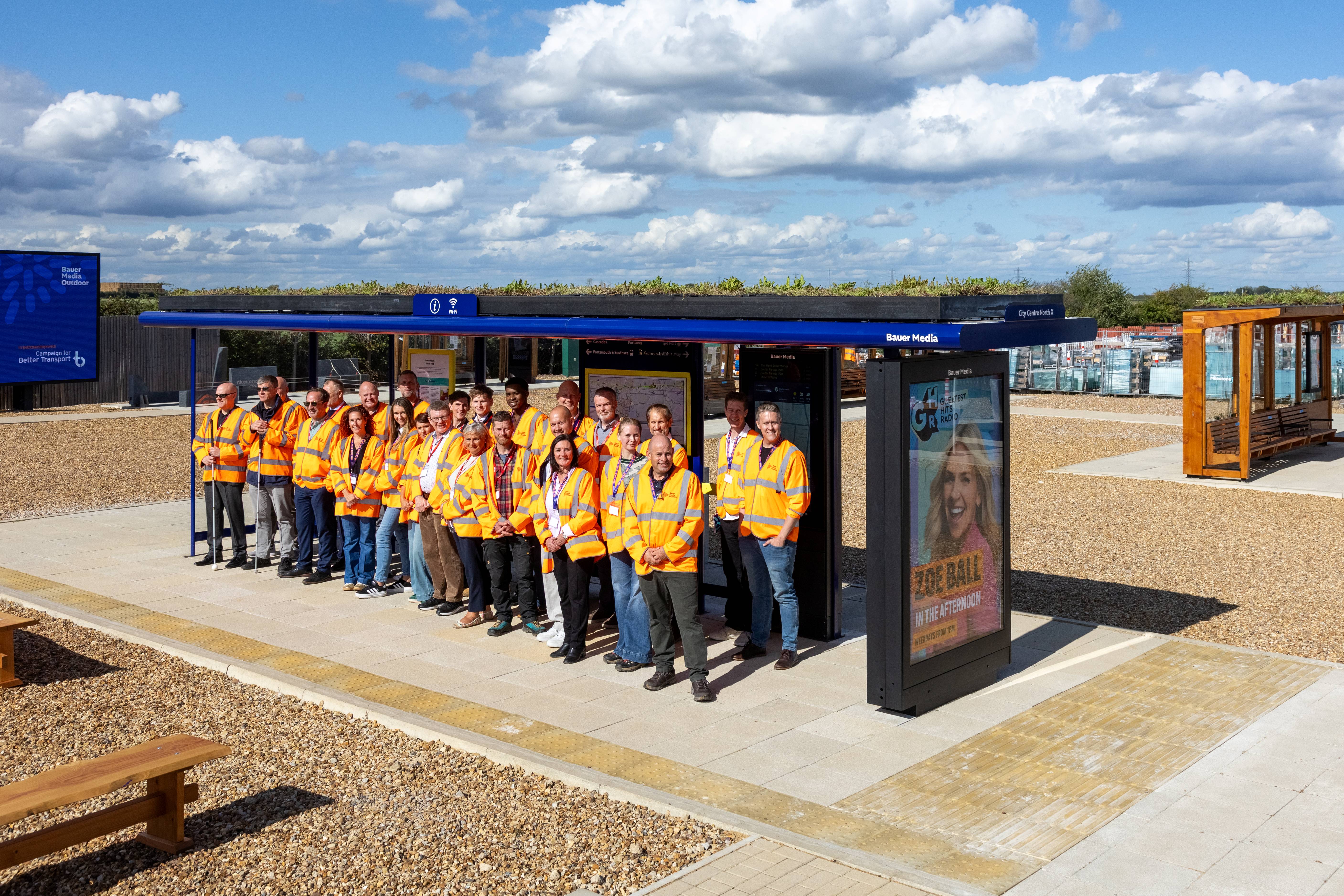 Group of Product Showcase attendees wearing high-visibility jackets standing beside a Bauer Media bus shelter with a Living Roof and digital advertising panel.