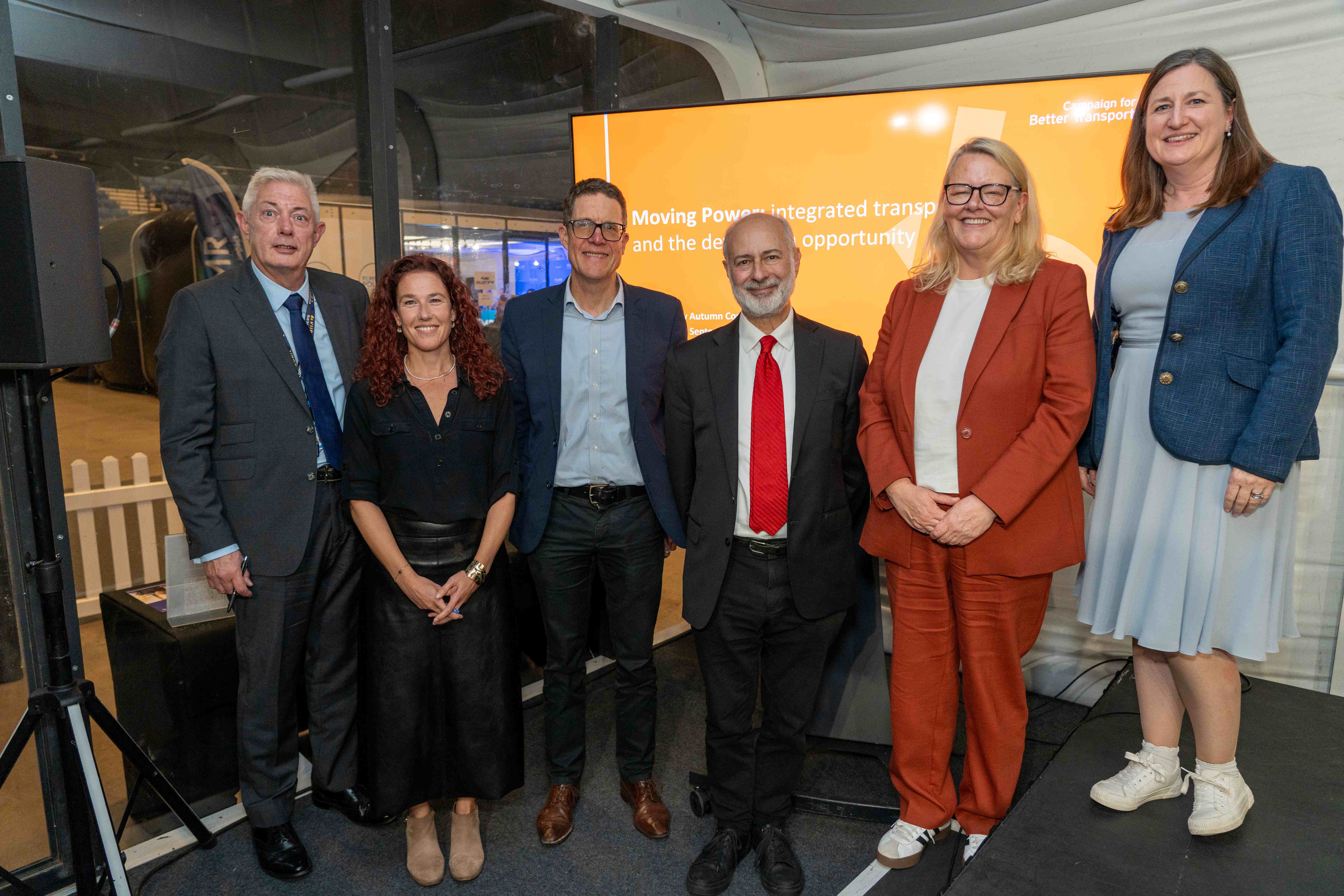Six people stand side by side, smiling at the camera at an indoor event, in front of an orange display about integrated transport. They wear a mix of suits and dresses, including a red tie, a rust-orange trouser suit and a blue blazer over a pale blue dress.