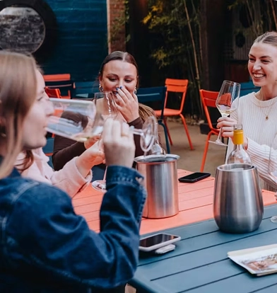Three women laugh and drink wine together at a colorful outdoor table on a patio.
