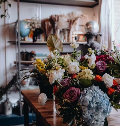 A lush floral arrangement with roses, hydrangeas, and mixed blooms on a wooden table in an eclectic, plant-filled shop.
