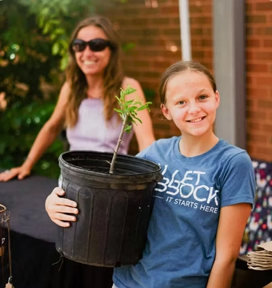 A smiling girl in a blue "Love Lubbock" T-shirt holds a potted seedling, with a woman smiling in the background.