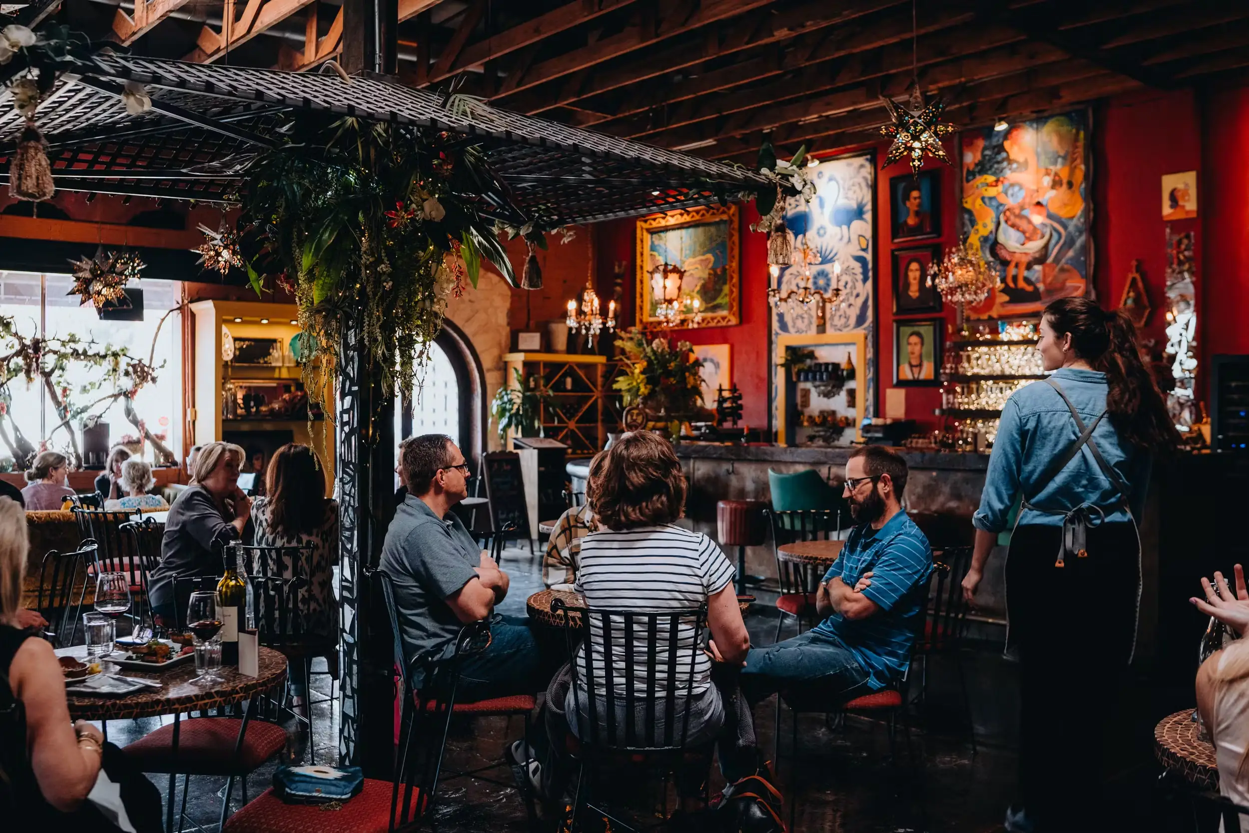 Diners chat at tables inside a warmly lit, eclectic Mexican-style restaurant with red walls, Frida Kahlo portraits, chandeliers, and hanging plants.