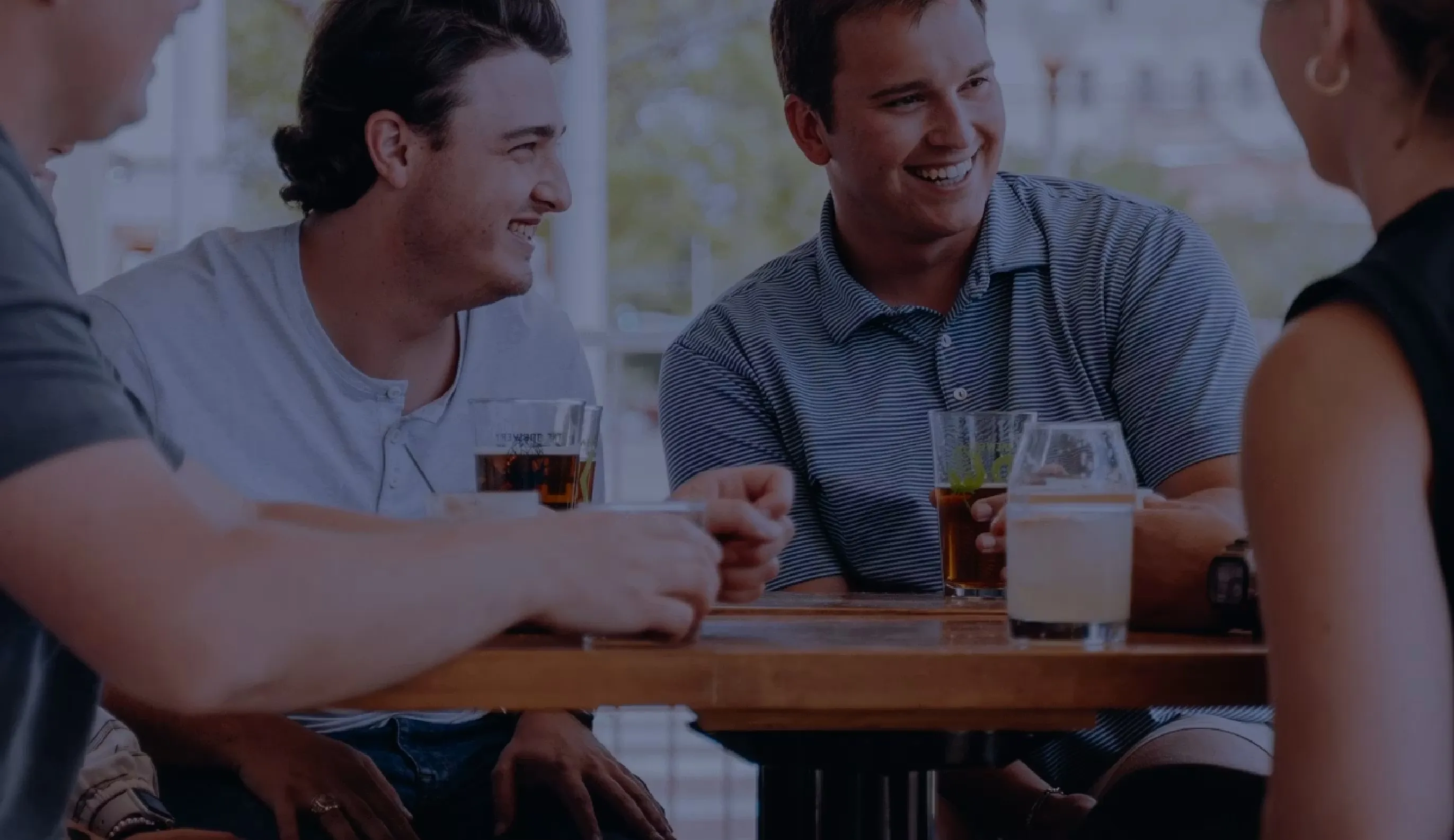 Group of people sitting at a table, smiling and holding glasses of beer or drinks in a social setting.