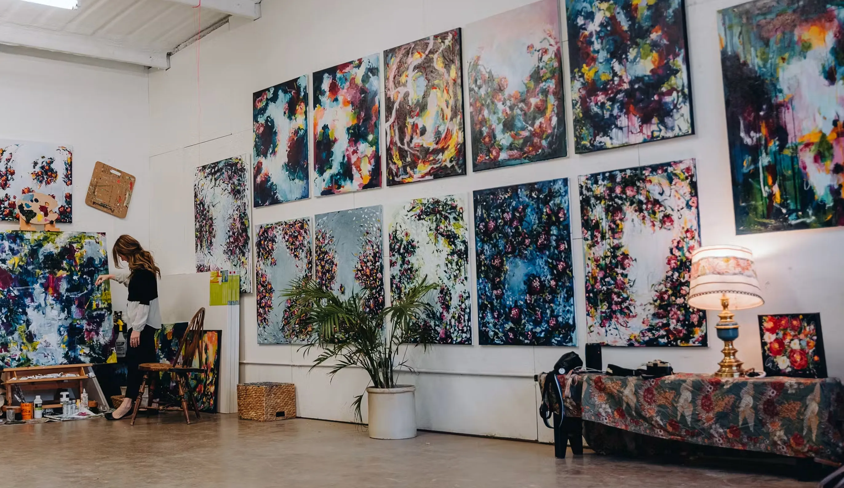 Artist in a studio surrounded by colorful abstract and floral paintings displayed on white walls.