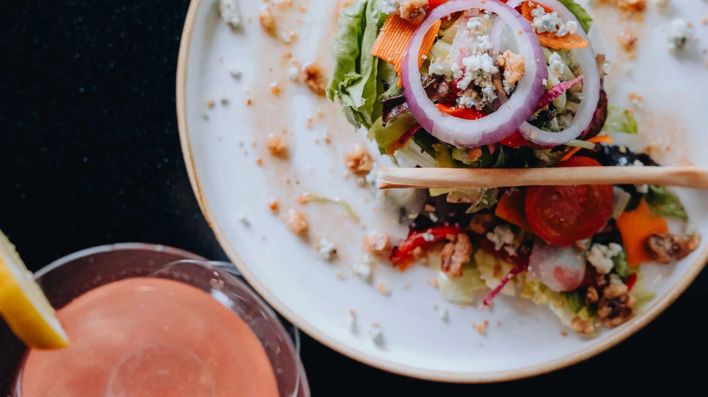 Plate with mixed salad topped with red onion rings, cherry tomato, and walnuts beside a pink drink with a lemon wedge.