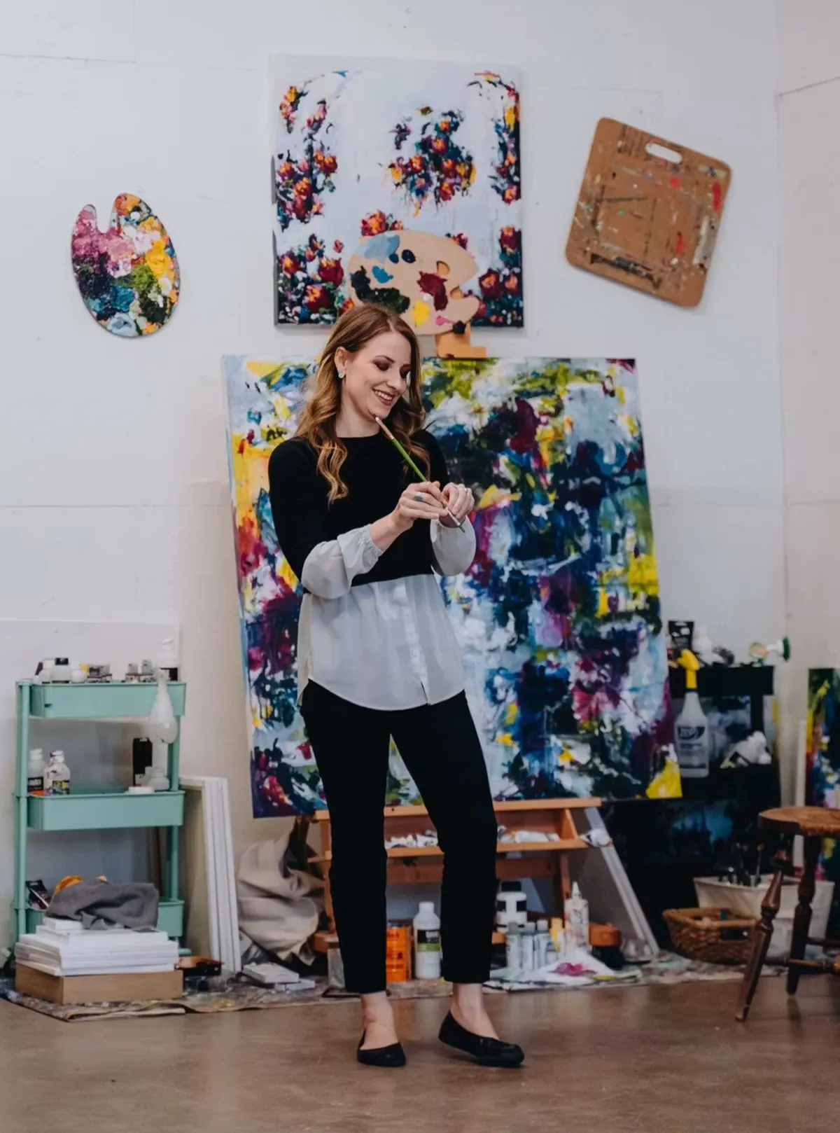 Woman holding a paintbrush in her art studio in front of colorful abstract paintings.