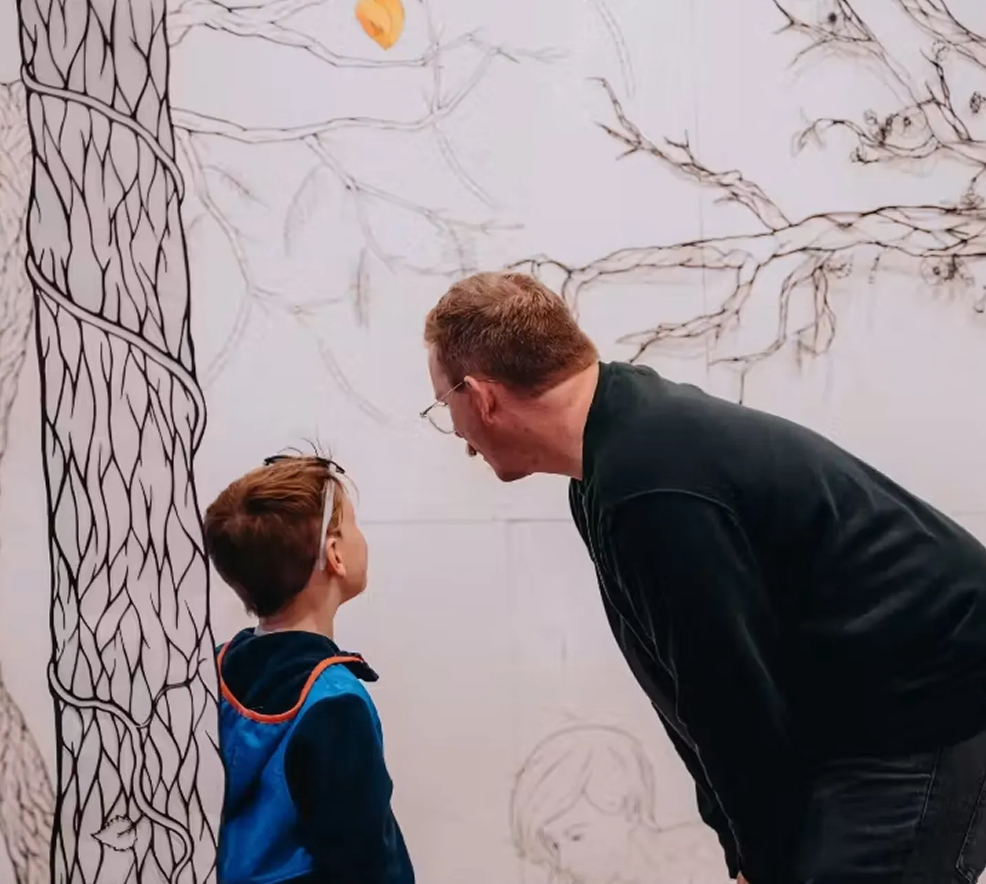 Adult and child talking in front of a tree-mural wall.