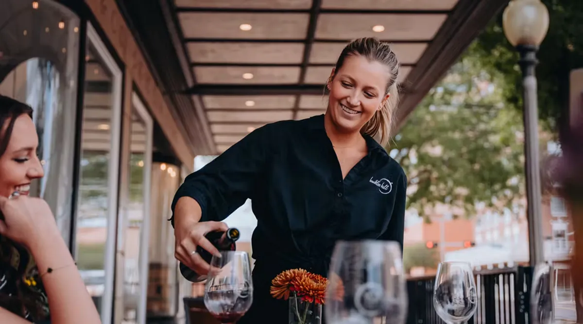 Server pouring red wine for a guest on a restaurant patio.