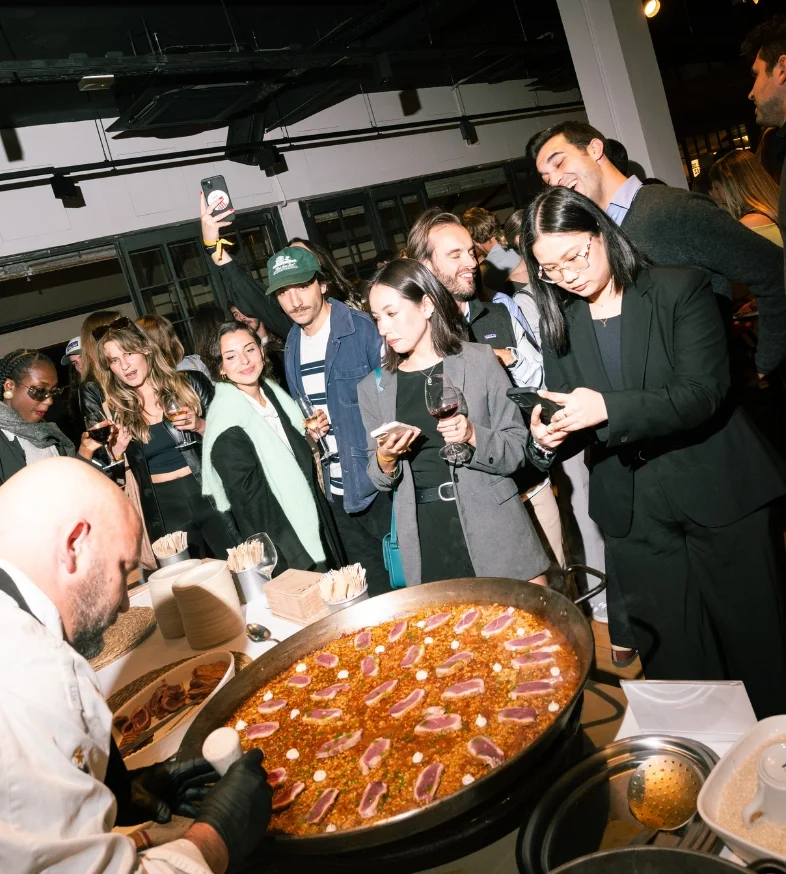 Un grupo de personas observa a un chef mientras cocina una gran paella en un evento social interior.