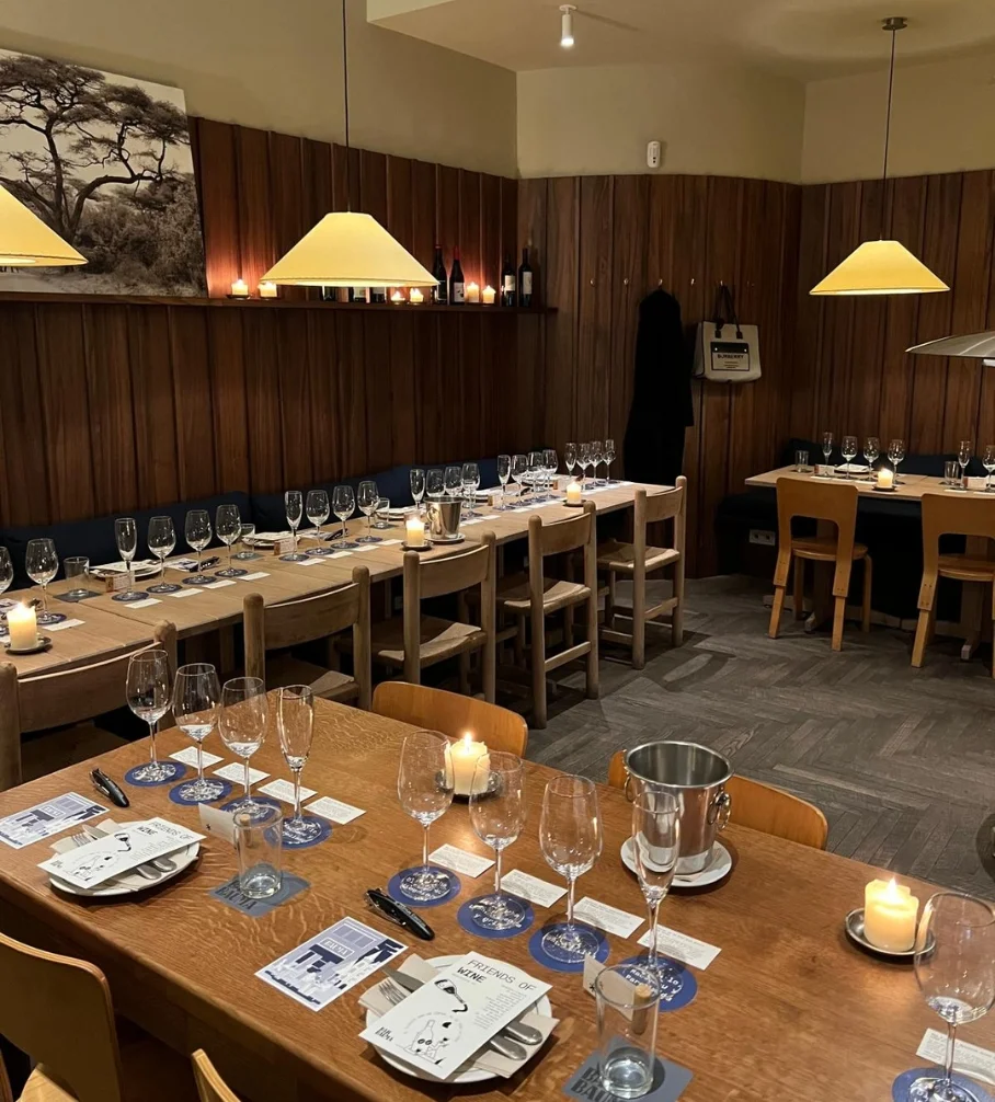 Mesa de madera en un restaurante con copas de vino, velas encendidas y cartas para cata de vinos, paredes revestidas en madera y luces colgantes cálidas.