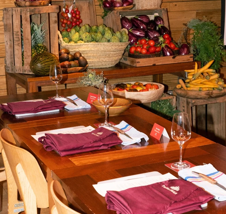 Mesa de madera con juegos de servilletas color vino, copas y cubiertos, rodeada de cajas y canastas con verduras frescas como jitomates, berenjenas, alcachofas, zanahorias y piñas.