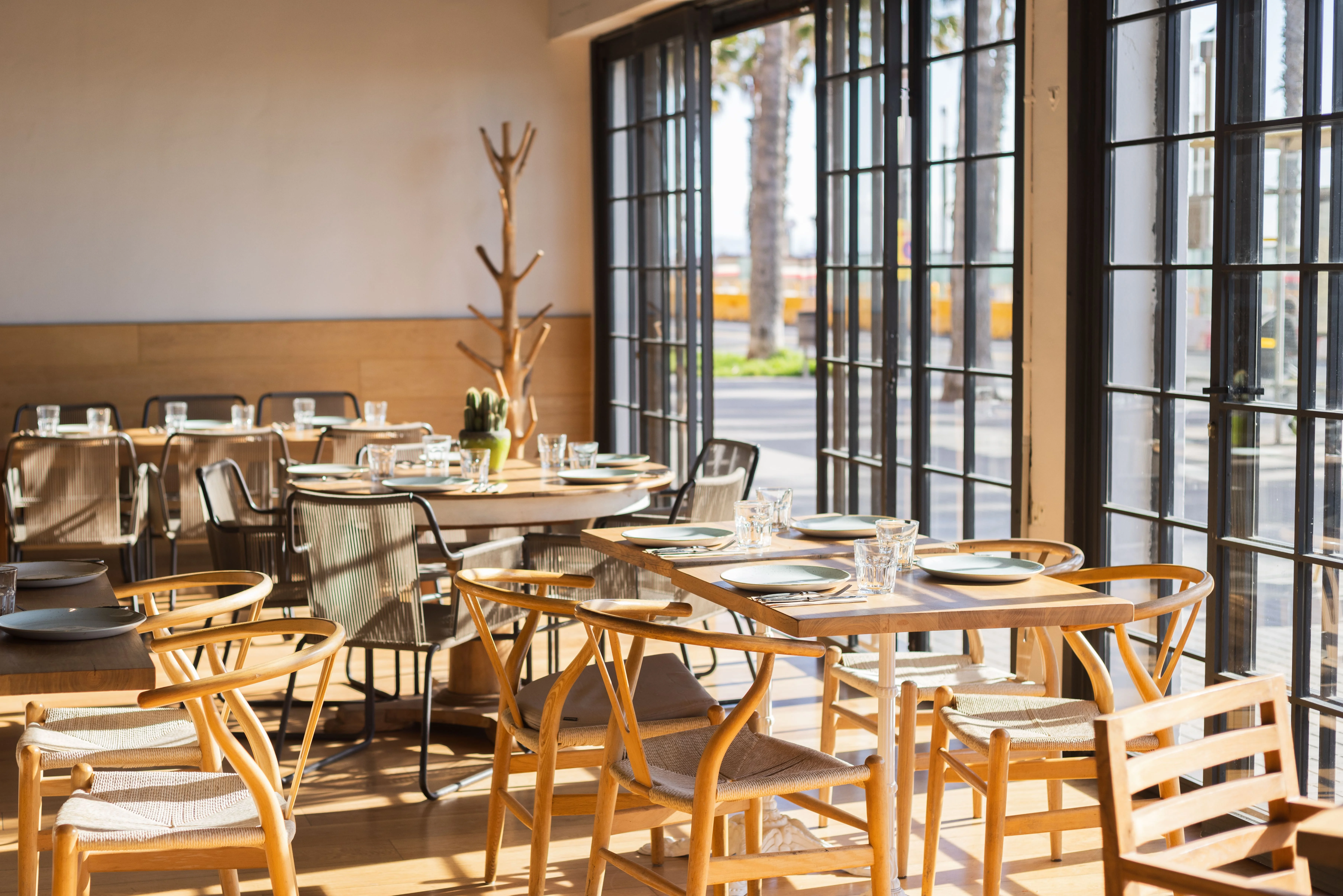Interior de restaurante luminoso con mesas de madera, sillas de diseño y grandes ventanas que dejan entrar la luz natural.