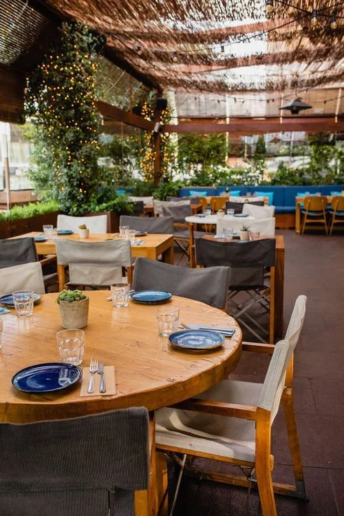 Restaurante con mesas de madera redondas y cuadradas, sillas de colores neutros, decoración con plantas, luces decorativas y techo de cañas.