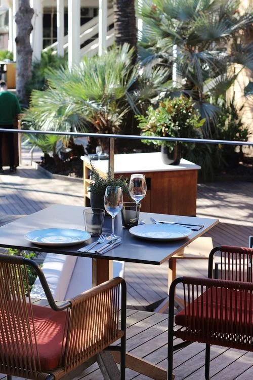 Mesa al aire libre con dos platos, copas de vino y cubiertos, rodeada de sillas con cojines rojos en un ambiente con vegetación y luz natural.