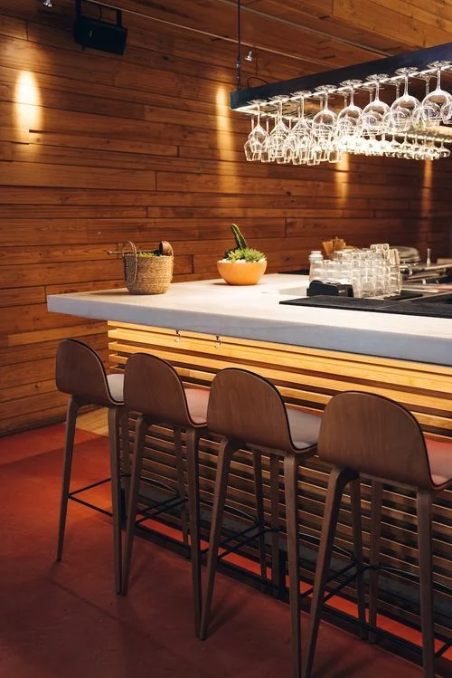 Barra de bar moderna con cuatro sillas altas de madera, copas colgadas boca abajo y una planta decorativa sobre la barra de mármol.