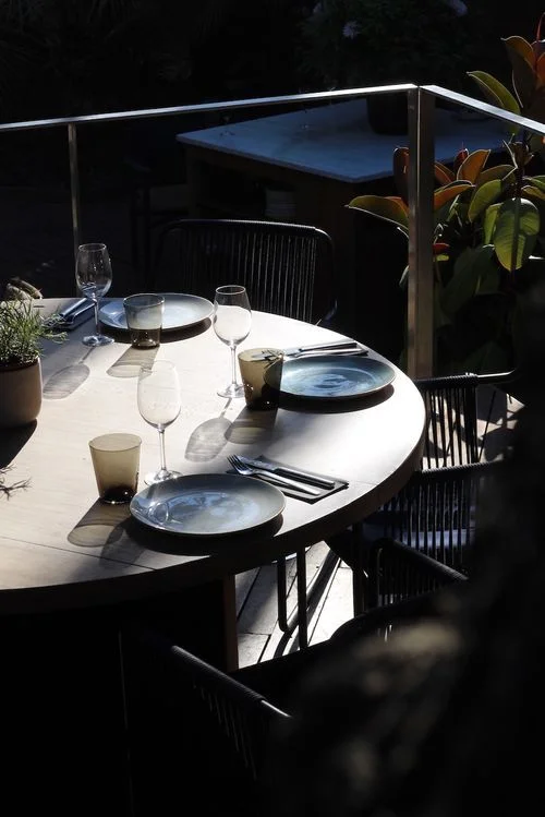 Mesa redonda al aire libre con platos, vasos y cubiertos, iluminada por luz natural con plantas alrededor.