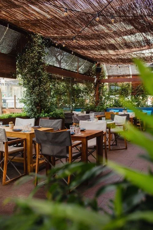 Área de restaurante al aire libre con mesas de madera, sillas con fundas grises y blancas, rodeado de plantas y techo de mimbre con luces colgantes.