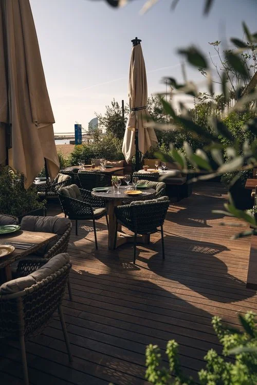 Terraza al aire libre con mesas con manteles, sillas tejidas y sombrillas cerradas bajo luz natural.