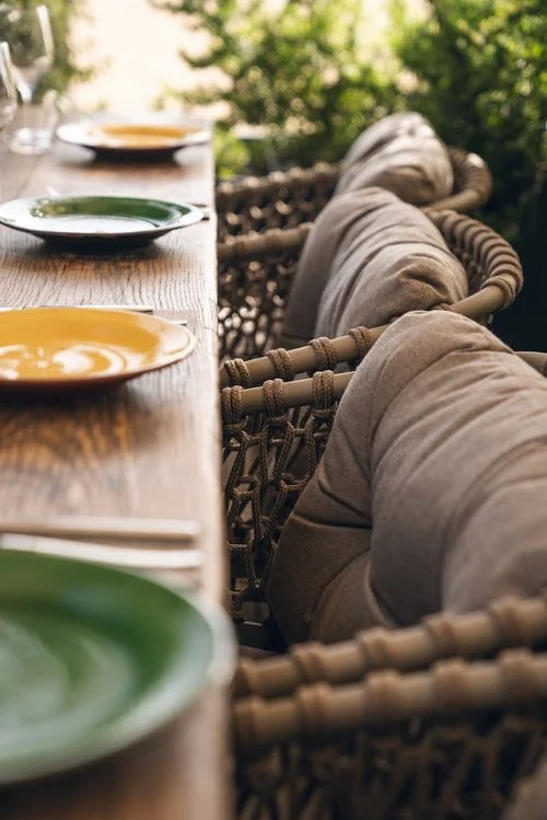 Mesa de madera con platos de colores y sillas de mimbre con cojines en un entorno exterior con vegetación.