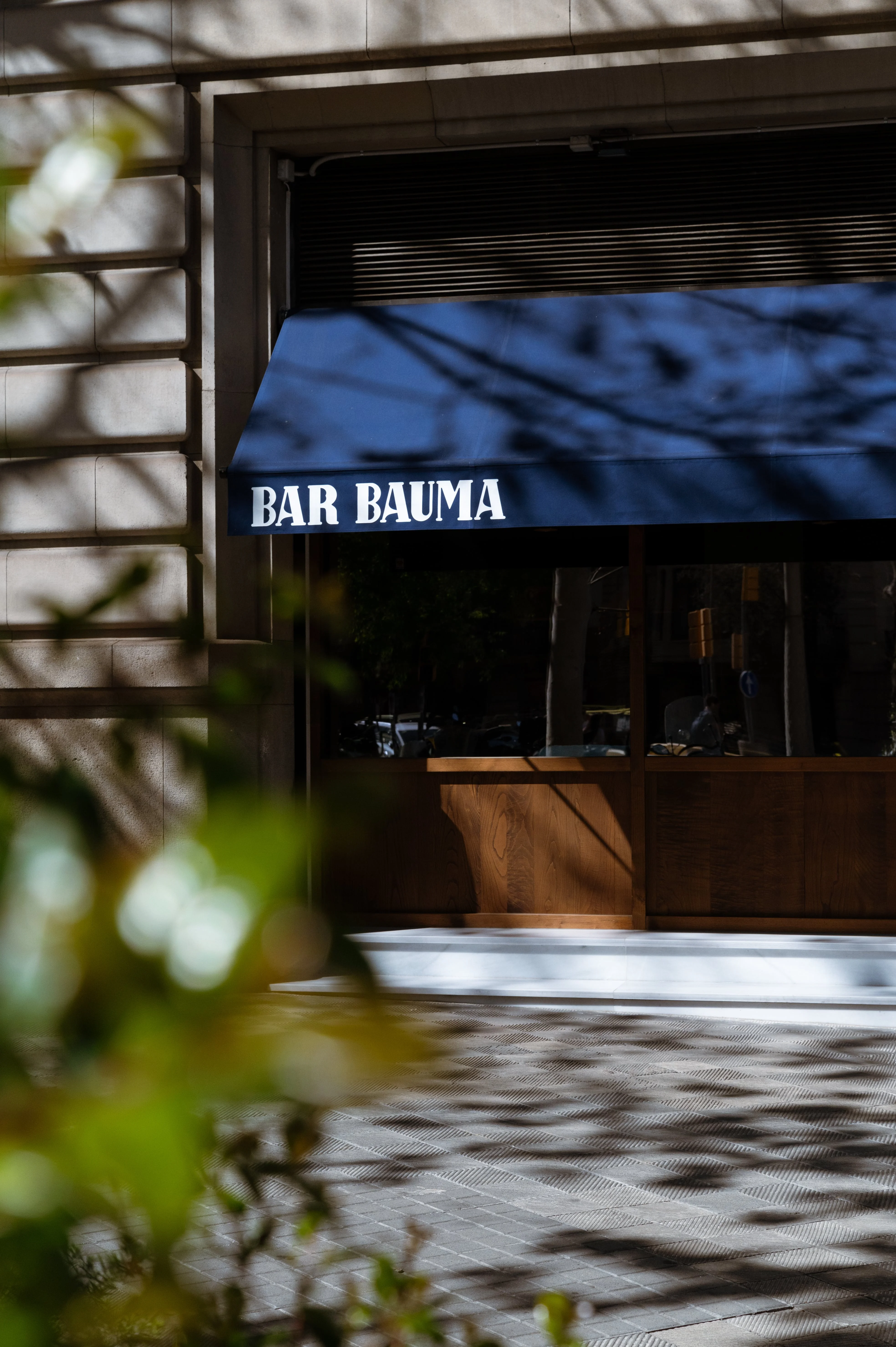 Entrada de Bar Bauma con toldo azul y fachada de piedra, con reflejos y sombras de árboles.