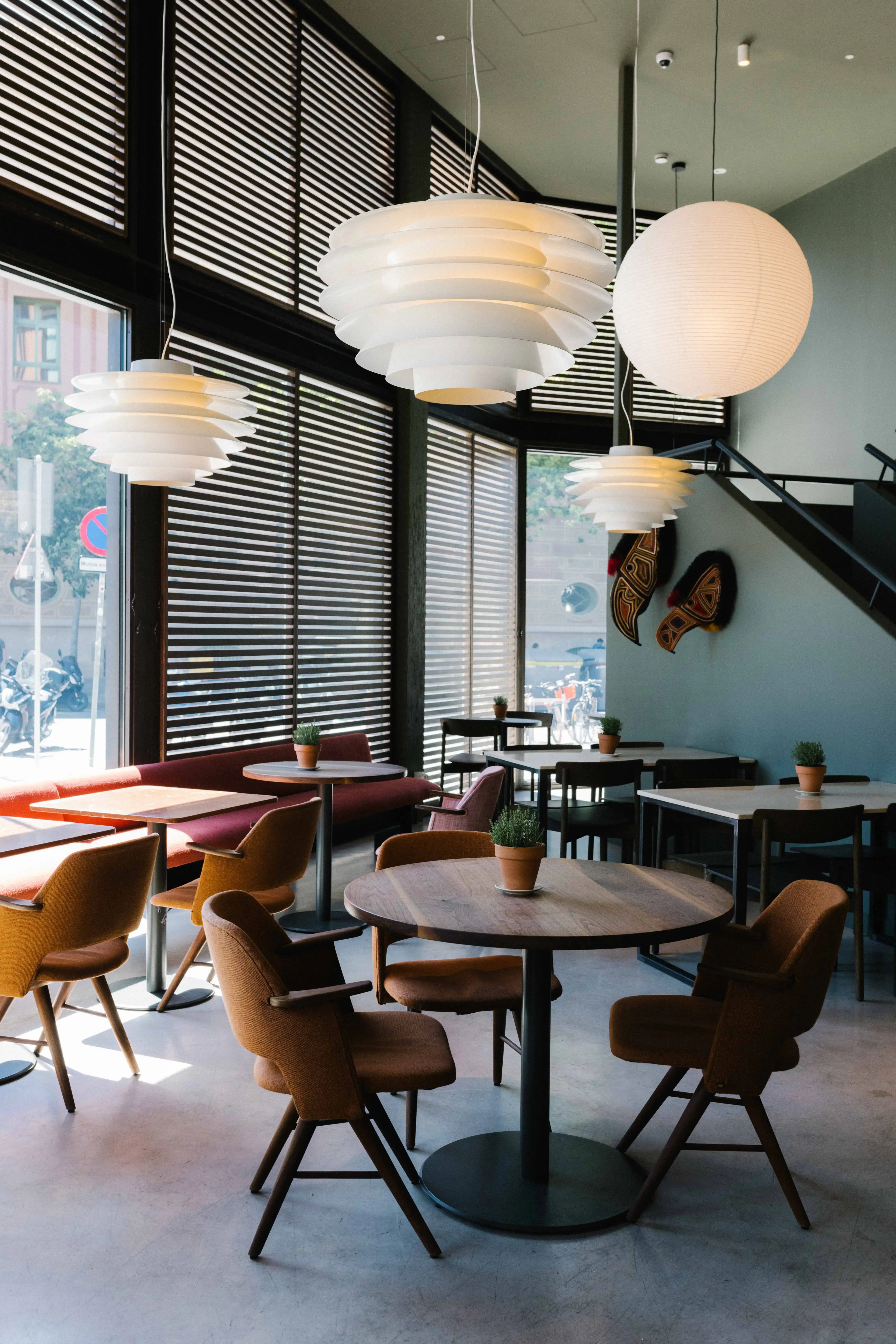 Intérieur de café moderne avec des tables rondes en bois, des chaises rembourrées brunes, de petites plantes en pot et des lampes suspendues blanches.