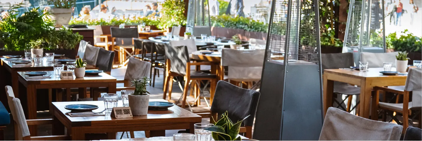 Tables en bois avec chaises et plantes dans un restaurant en plein air avec chauffage et lumière naturelle.