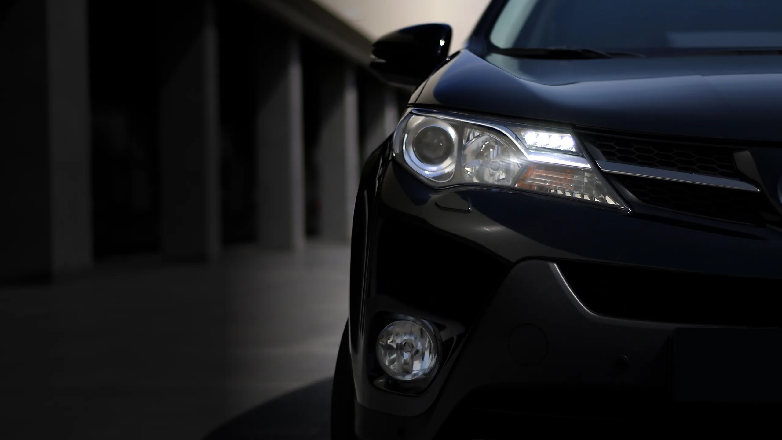 Close-up of the front right side of a black car showing the headlight, fog light, and side mirror in a dimly lit urban setting.
