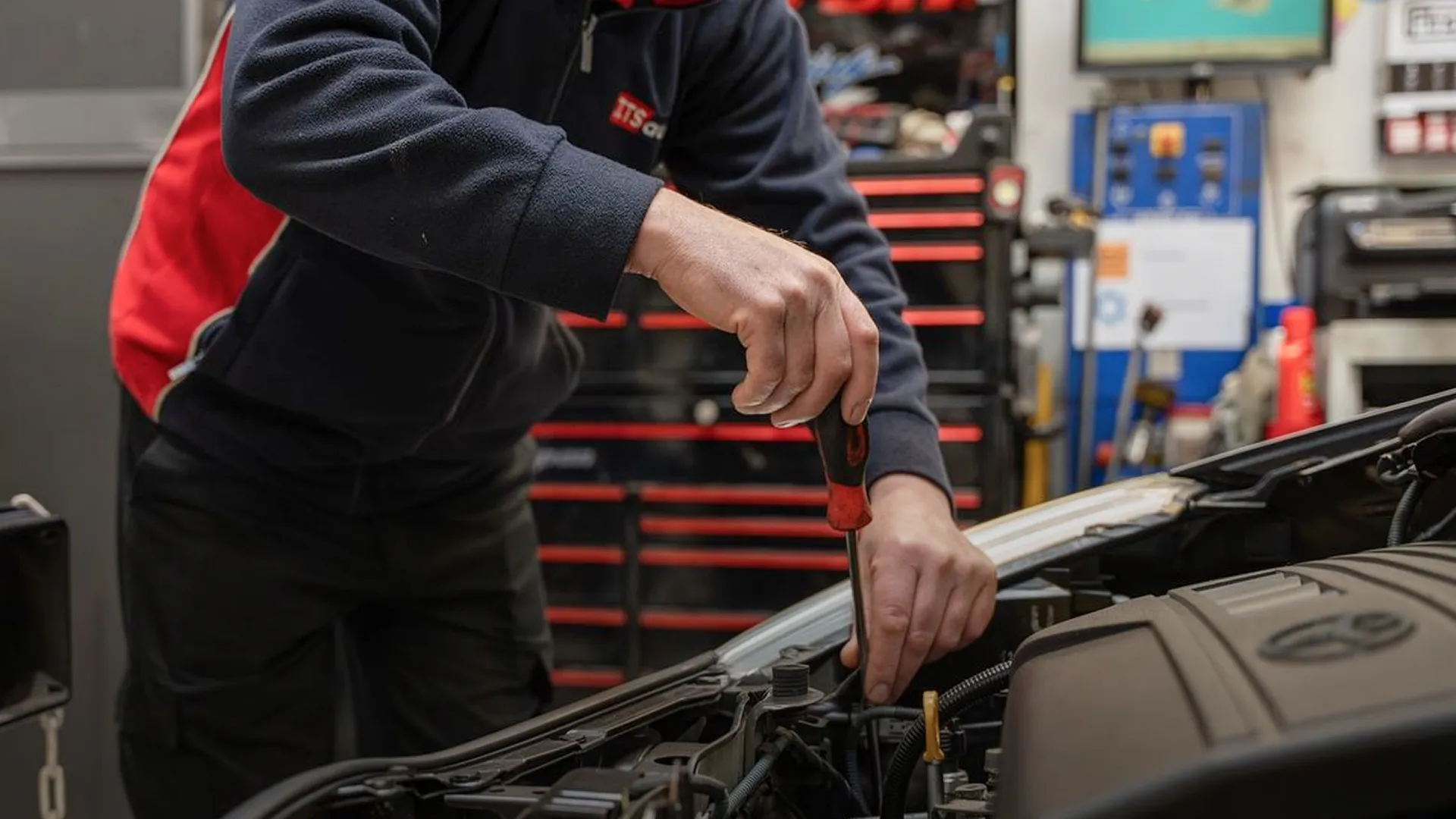 Mechanic in a navy and red jacket using a screwdriver to work on a car engine.