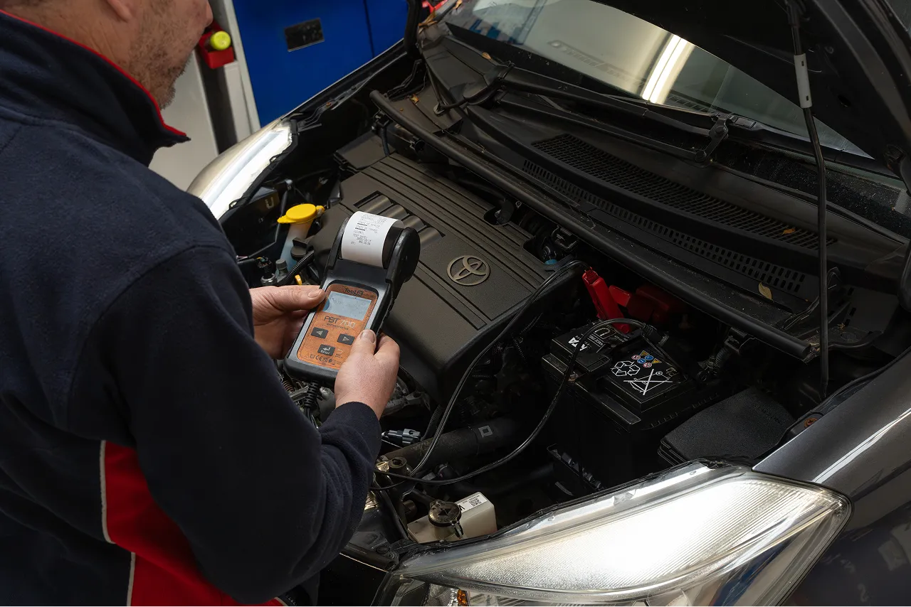 Person using a diagnostic tool to check the battery in a car engine compartment with a Toyota logo on the engine cover.