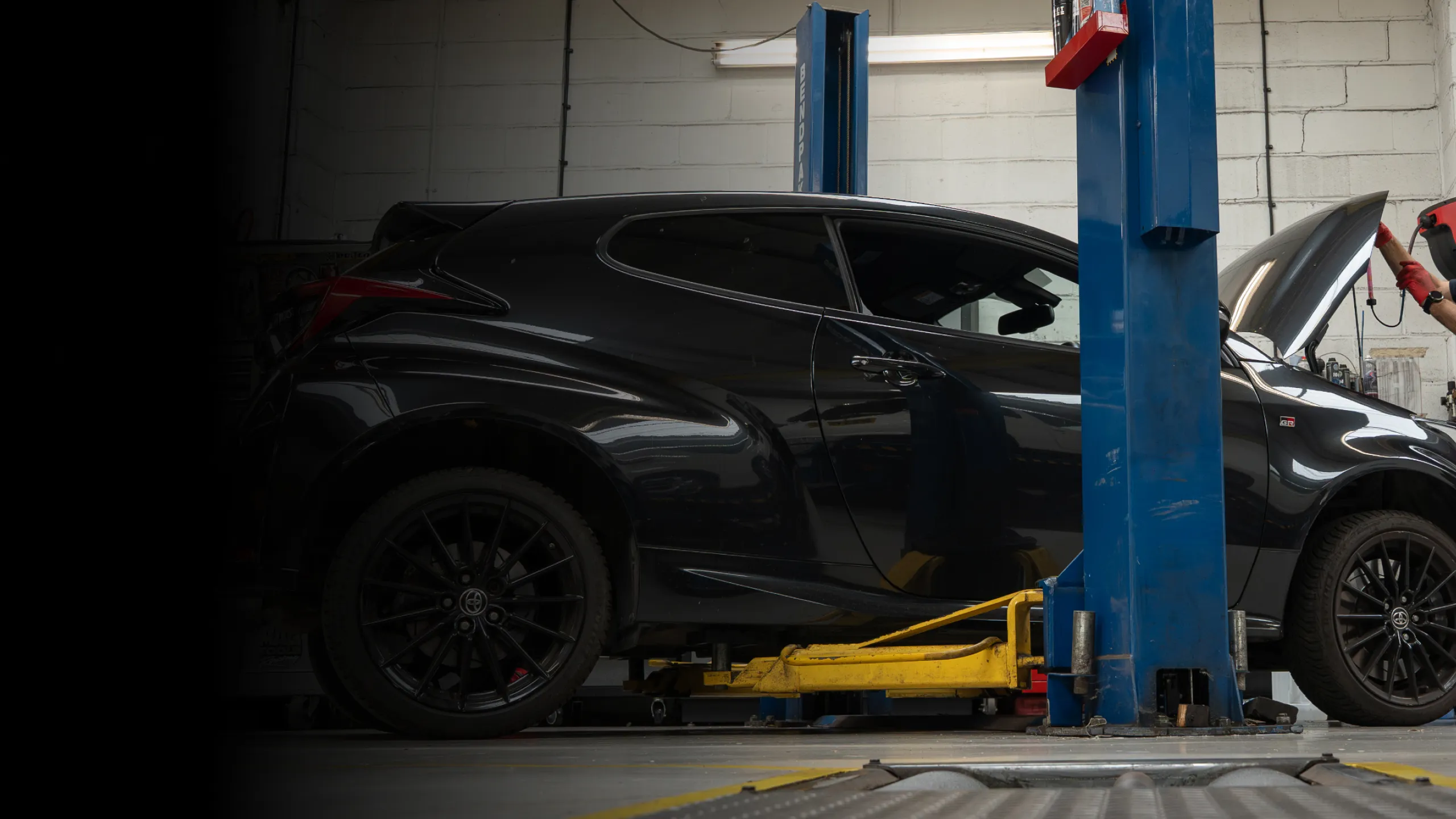 Black sports car on a yellow hydraulic lift inside a garage with a person inspecting the open hood.