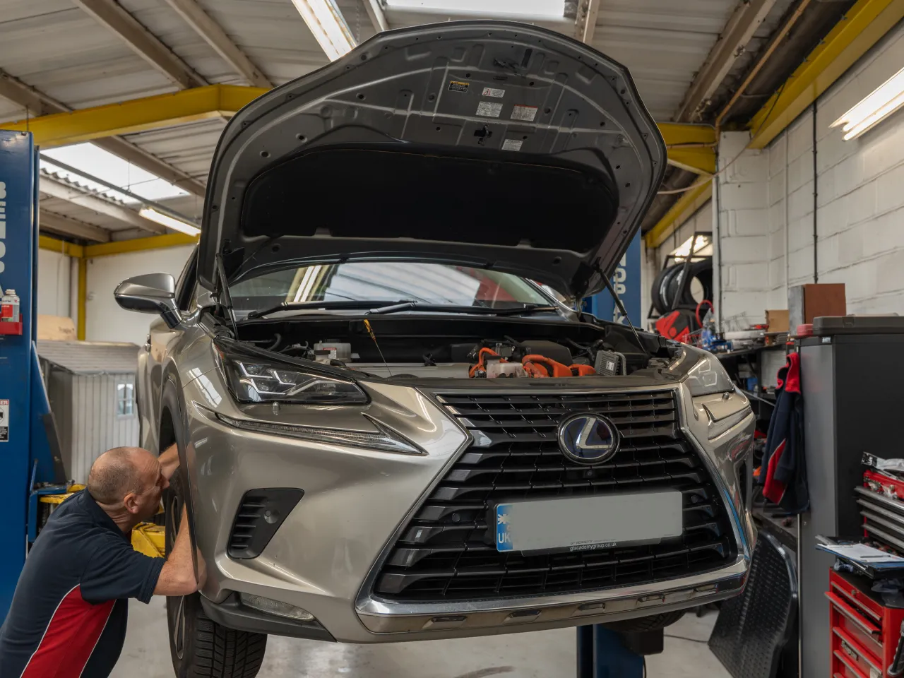 Mechanic inspecting the front wheel of a lifted silver Lexus SUV with its hood open in a garage.