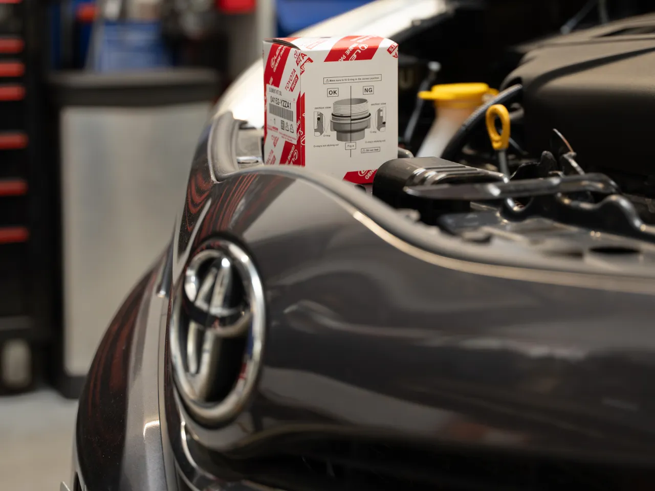 Close-up of an open car hood with a Toyota emblem on front and a Toyota-branded car part box placed on the engine.